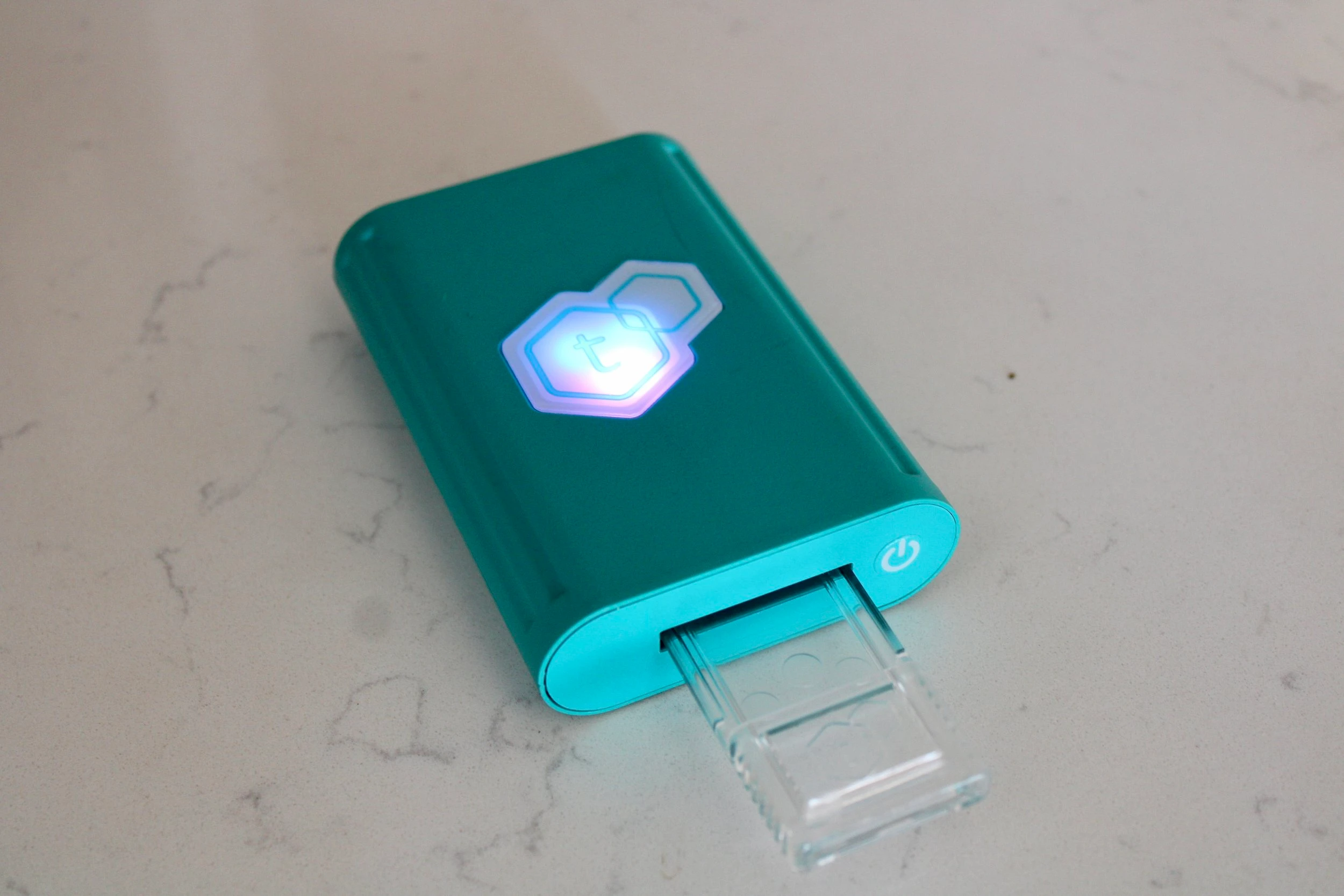 A turquoise tcheck potency tester turned on and lit up, resting on a marble countertop. 