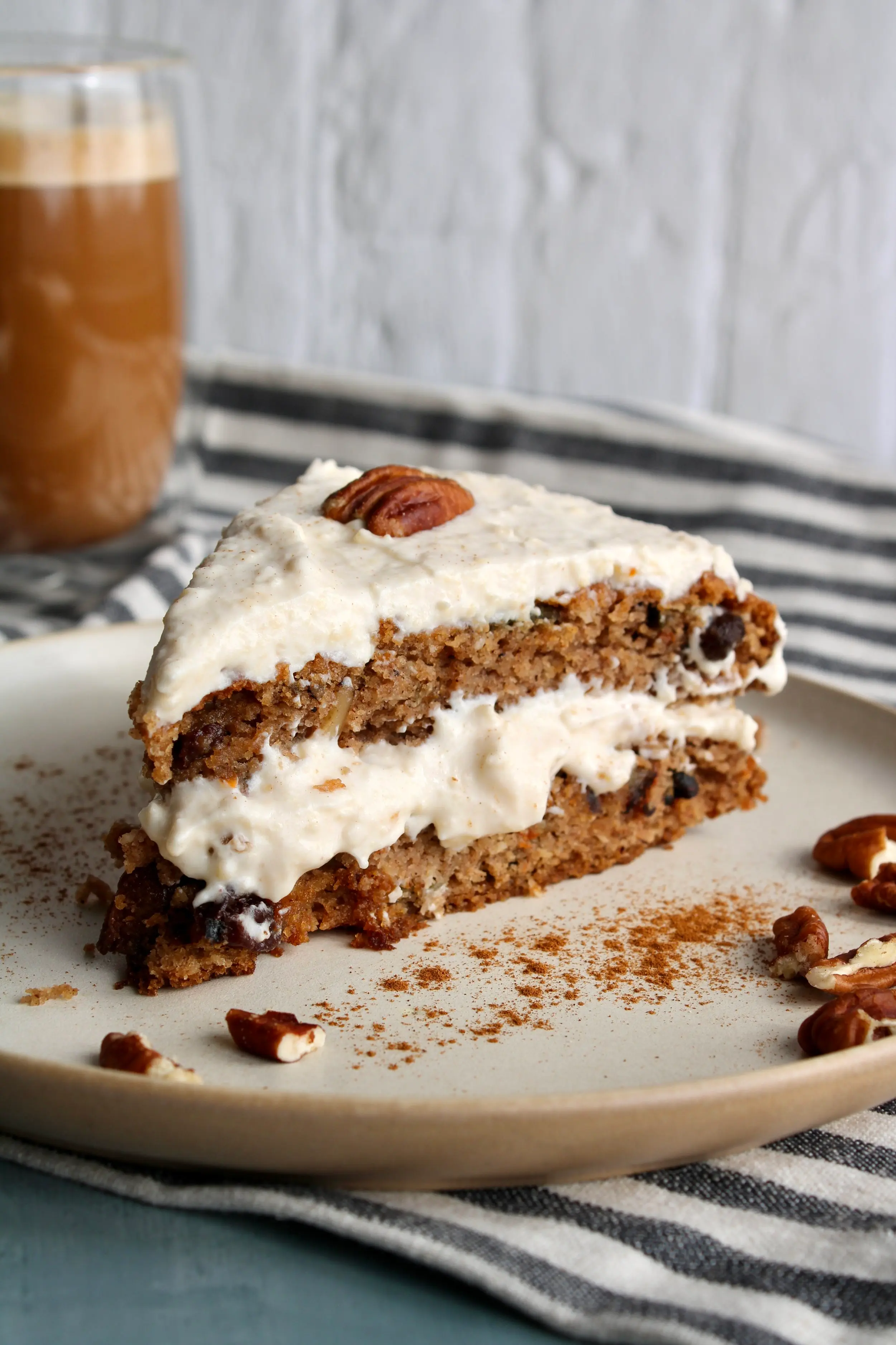 A cream plate of a large slice of carrot cake with creamy layers of frosting in the middle and on top. The cake is garnished with a pecan.