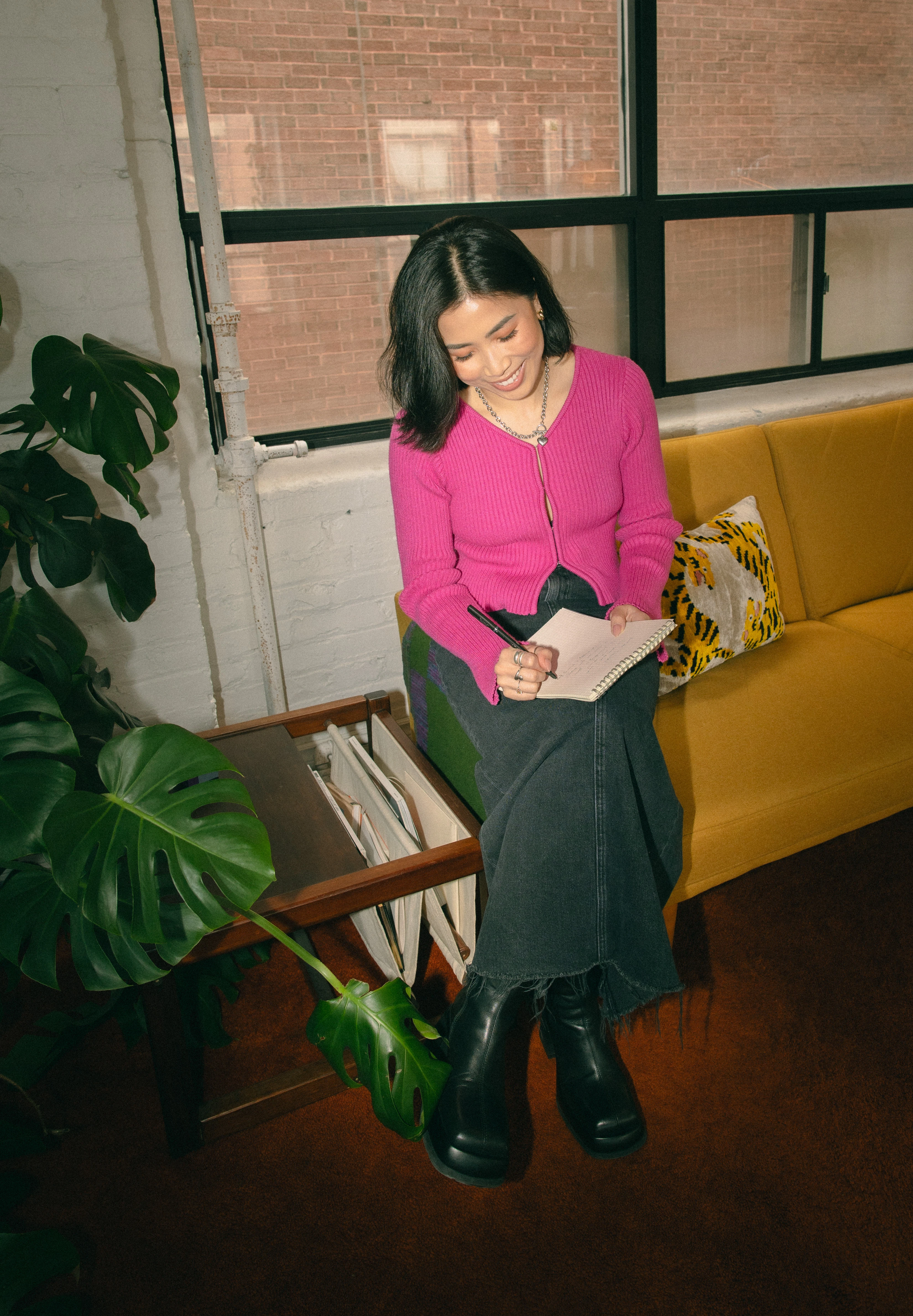 Asian woman in a pink cardigan sitting on a yellow sofa journalling.