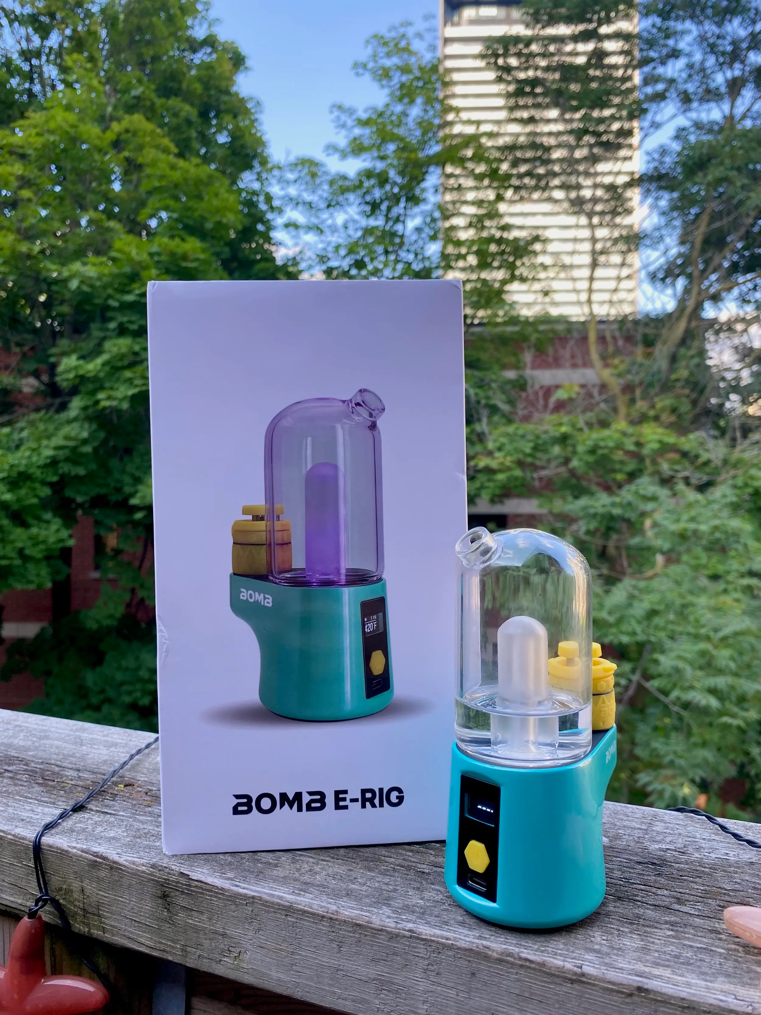 A teal electronic dab rig by the brand BOMB E-Rig which sits on the wooden edge of a fire escape right next to the white box it came in.