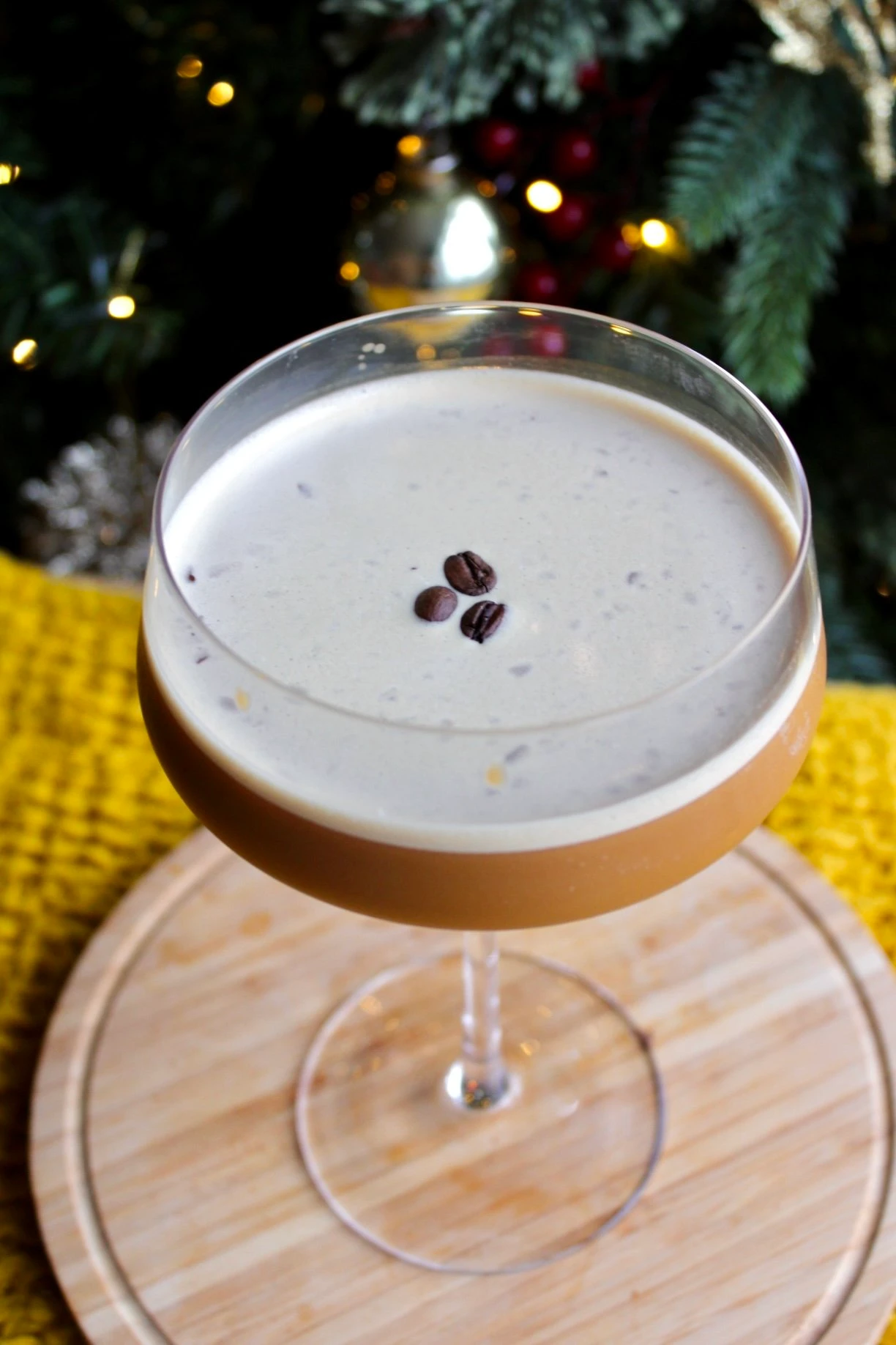 A martini glass holding a cannabis infused espresso martini mocktail, rest8ing on a wooden coaster in front of a Christmas tree.