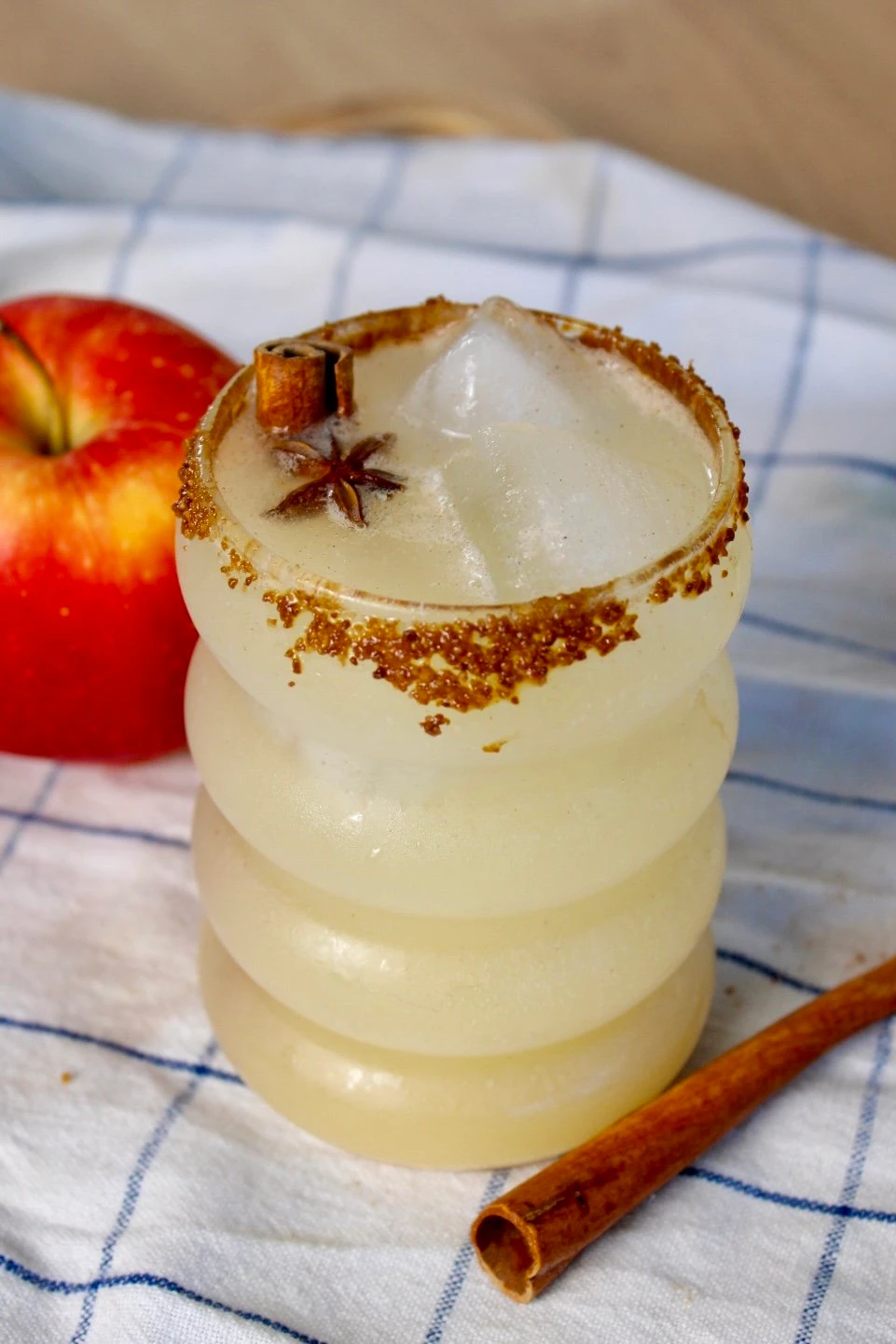 A bubble glass which holds an apple cider mocktail and rests on a checkered blue and white blanket. In front of it sits a cinnamon stick and behind it, a red apple.