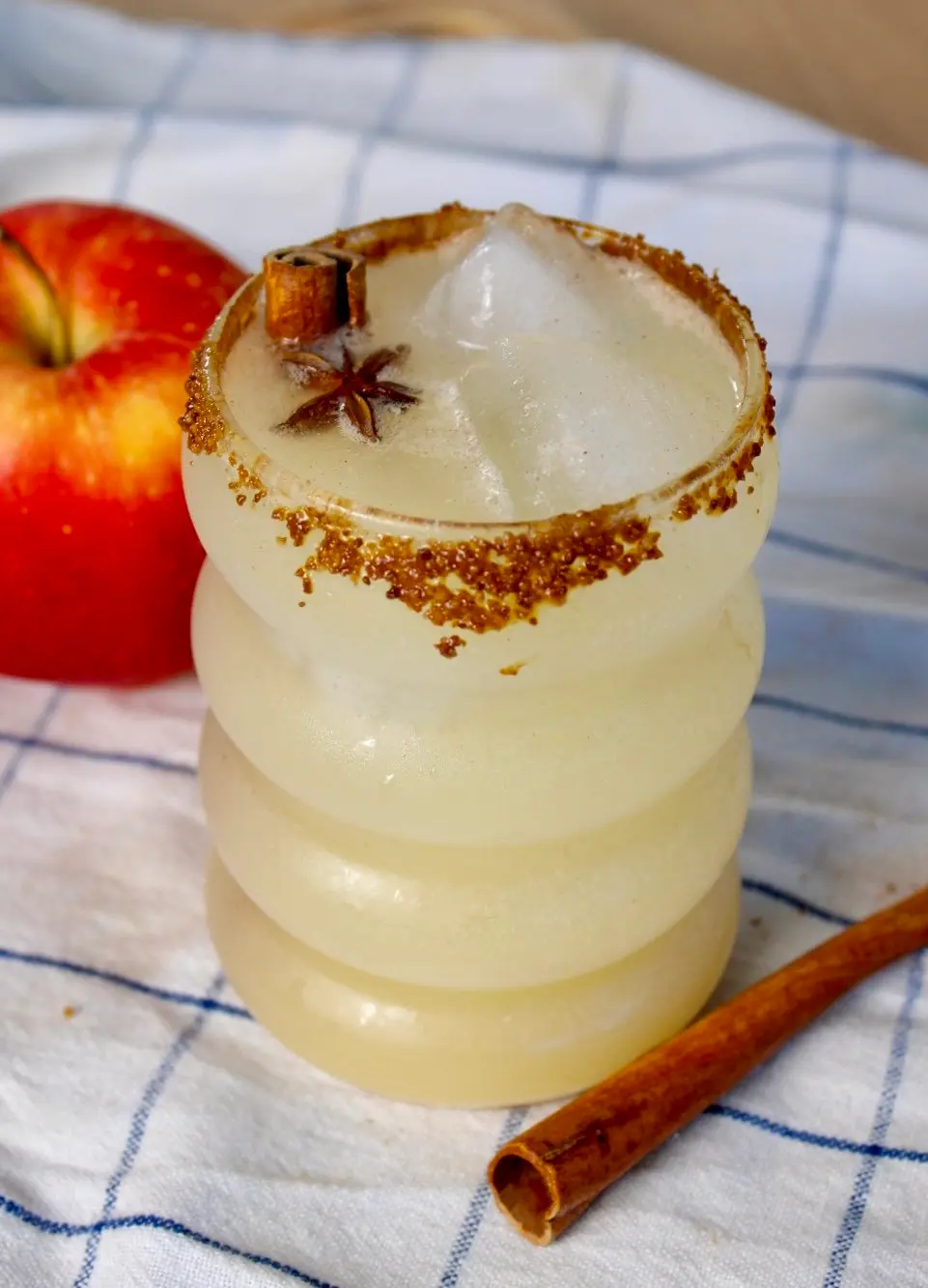 a bubble looking glass filled to the brim with a mild yellow drink, garnished with a star anise, while a cinnamon stick rests on a checkered cloth right next to it.