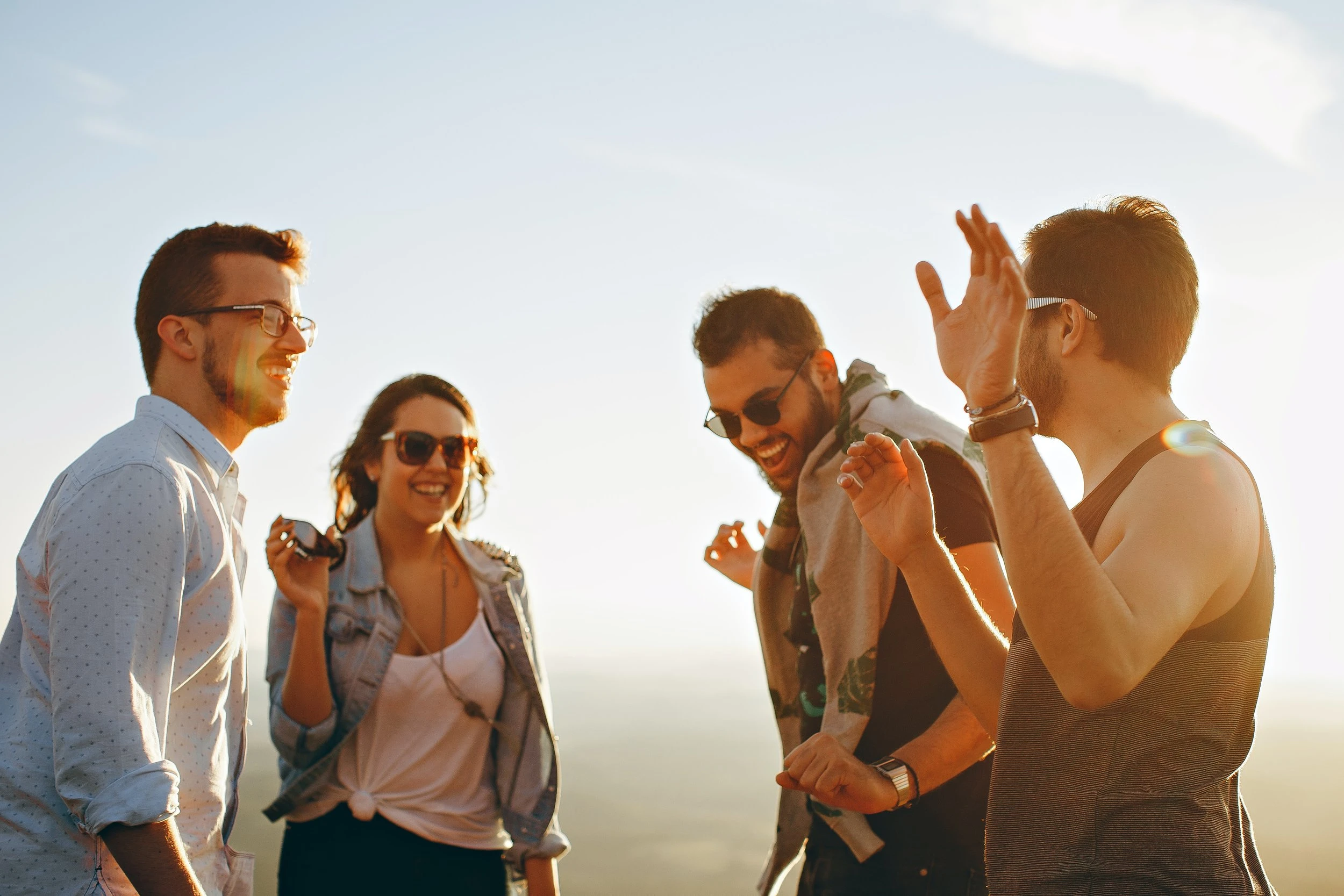 A group of four people, all wearing sunglasses, laughing with each other and engaging in a conversation with a blue sky in the background. 