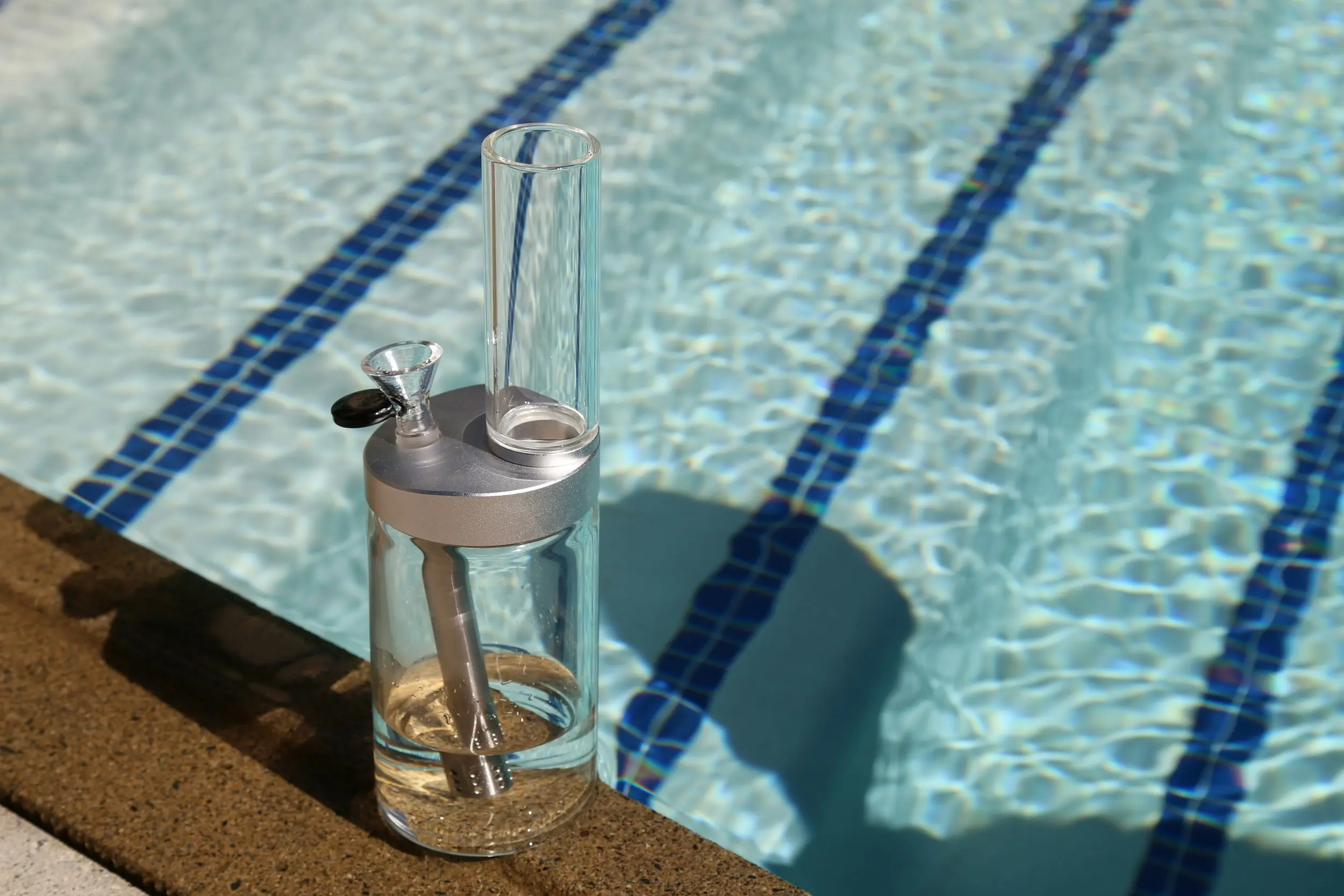 A glass bong is sitting at the very edge of a recreational swimming pool with teal and dark blue tiled lines along the bottom of it. 