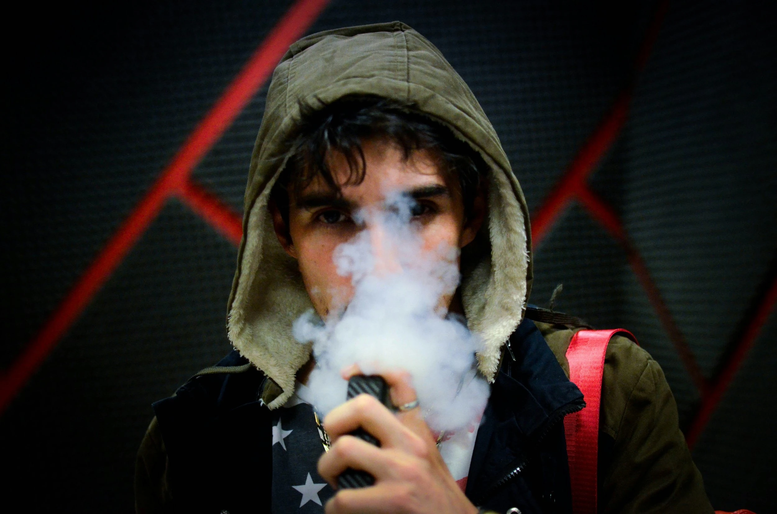 A boy holding a dried herb vaporizer and exhaling a large quantity of vapour with a green hood from his hoodie on.