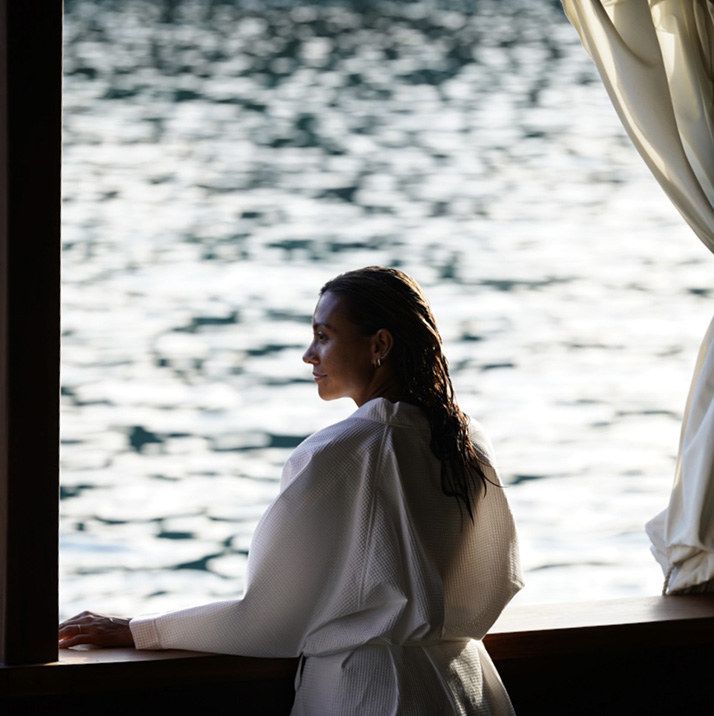 A guest in a white robe looks out of a yacht cabin window at sunlit water.