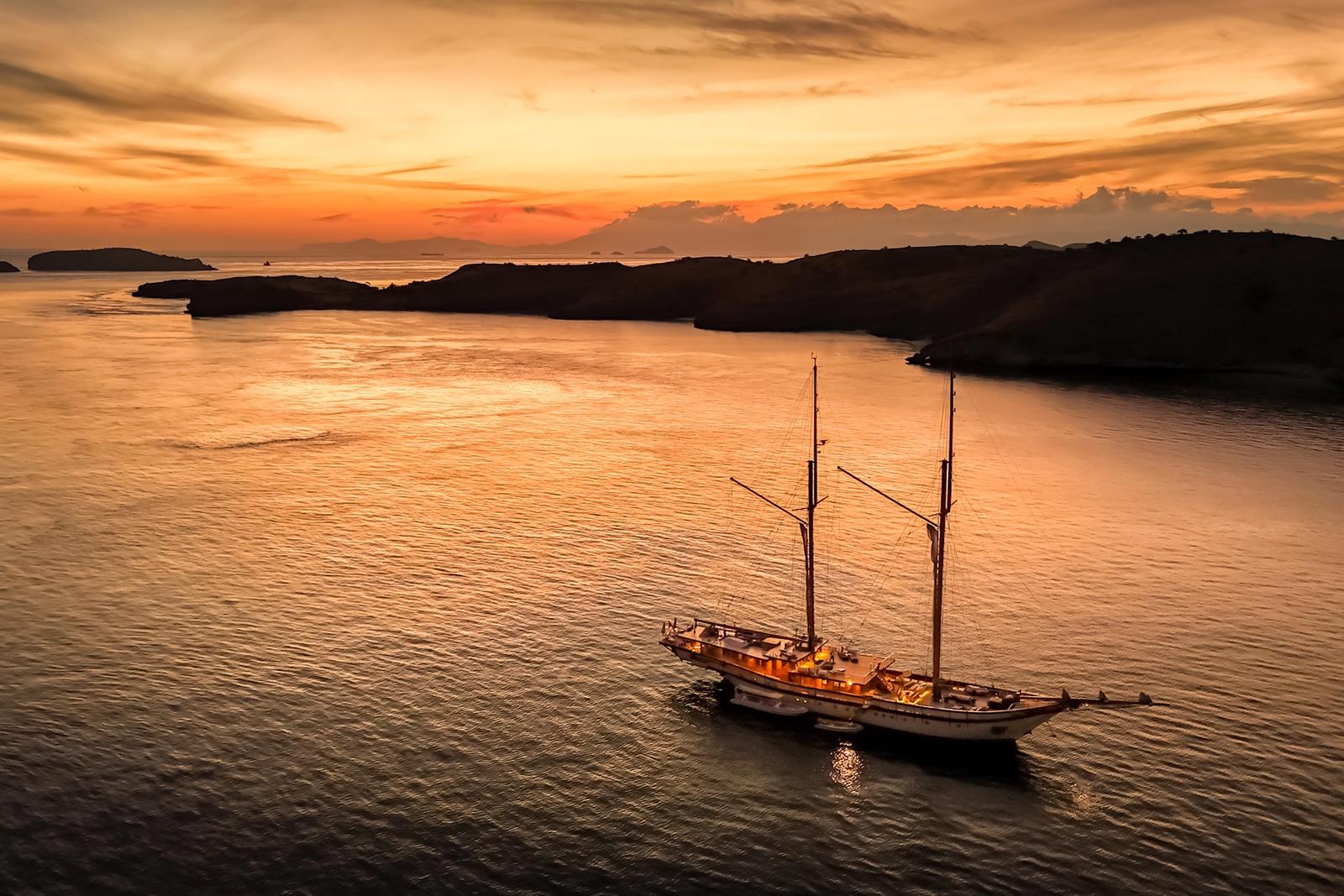 Aerial view of a phinisi anchored alone in a calm bay in Komodo as the sun sets.