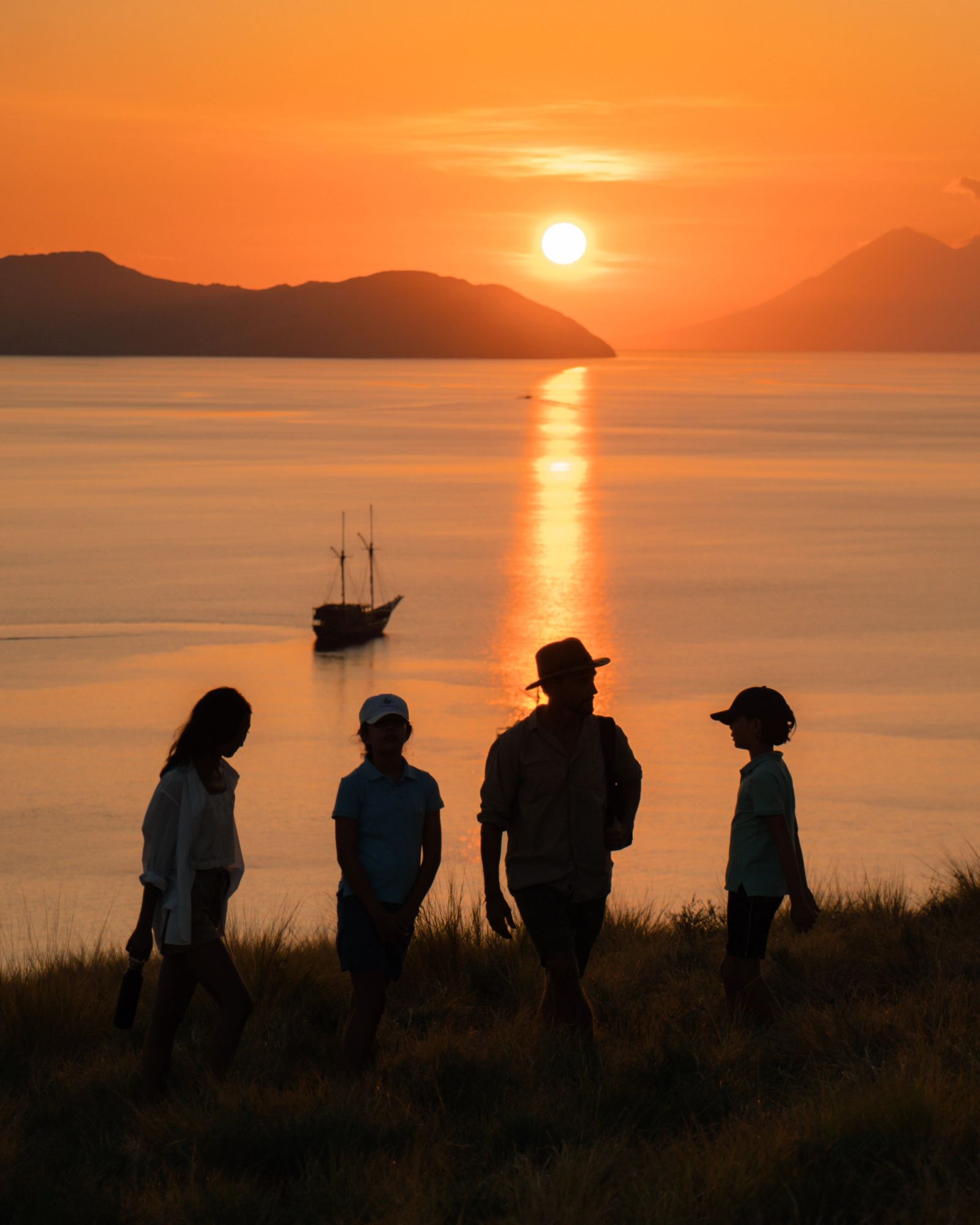 Guests silhouetted on a hilltop watching the sunset with a phinisi anchored below.