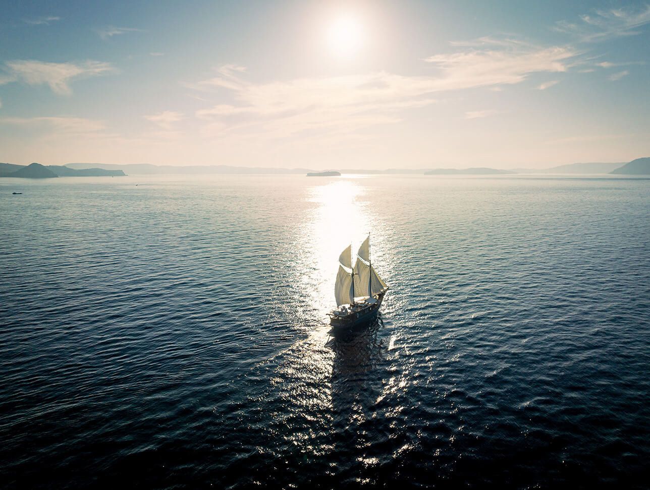A phinisi sailing toward a low sun that glitters on the water.