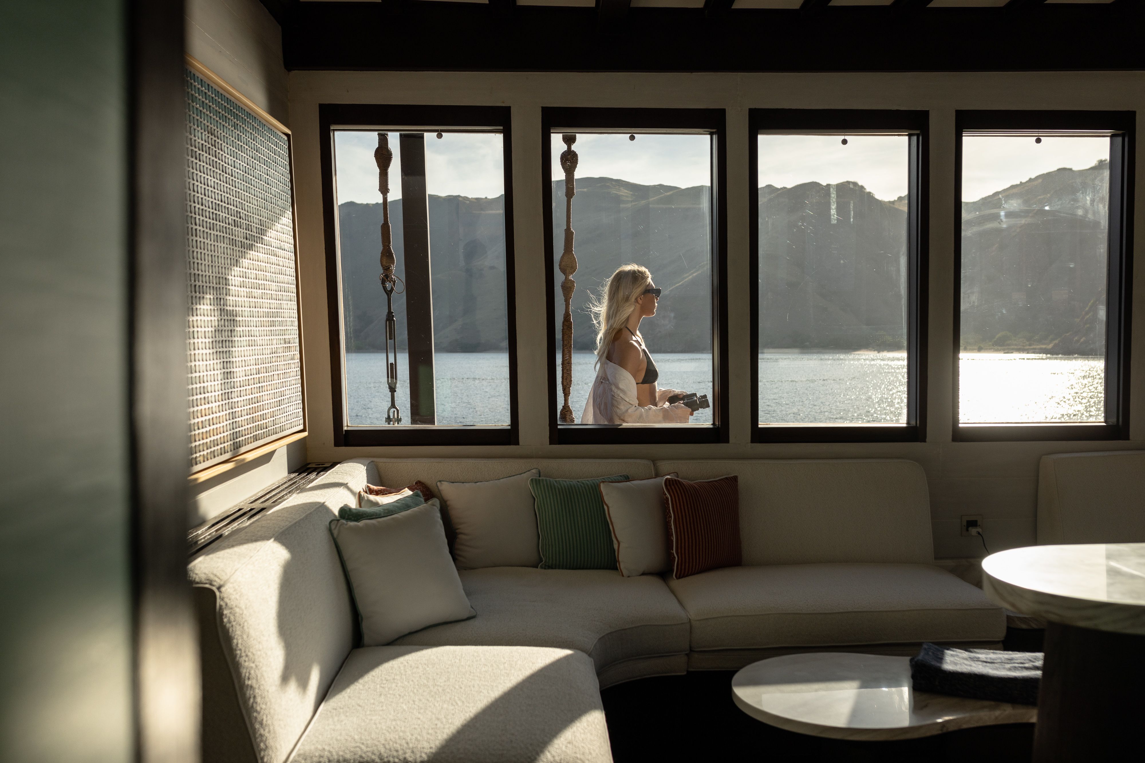 A guest stands at the windows of Vela's main saloon, with Komodo's hills beyond the glass.