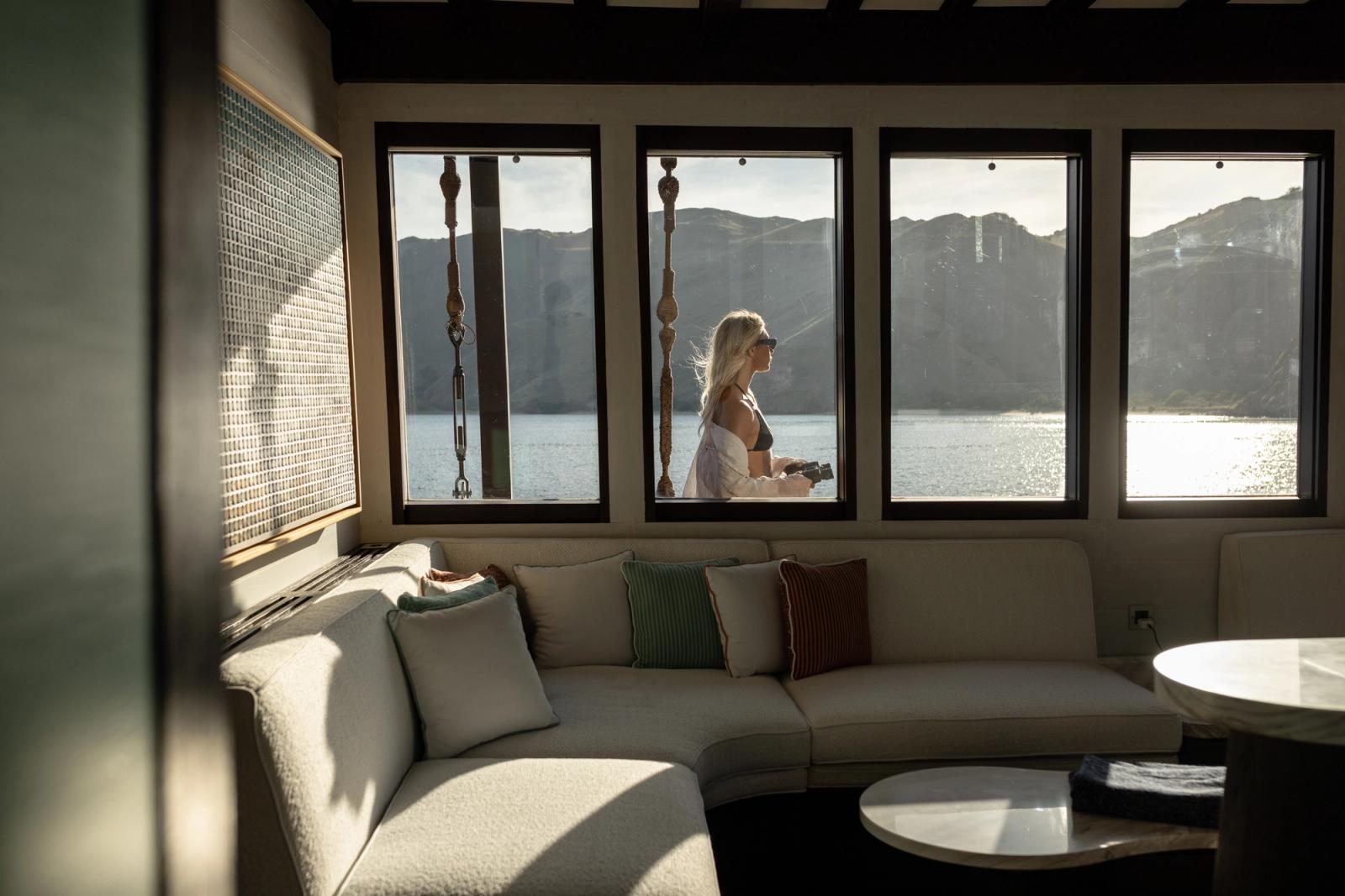 A guest stands at the windows of Vela's main saloon, with Komodo's hills beyond the glass.