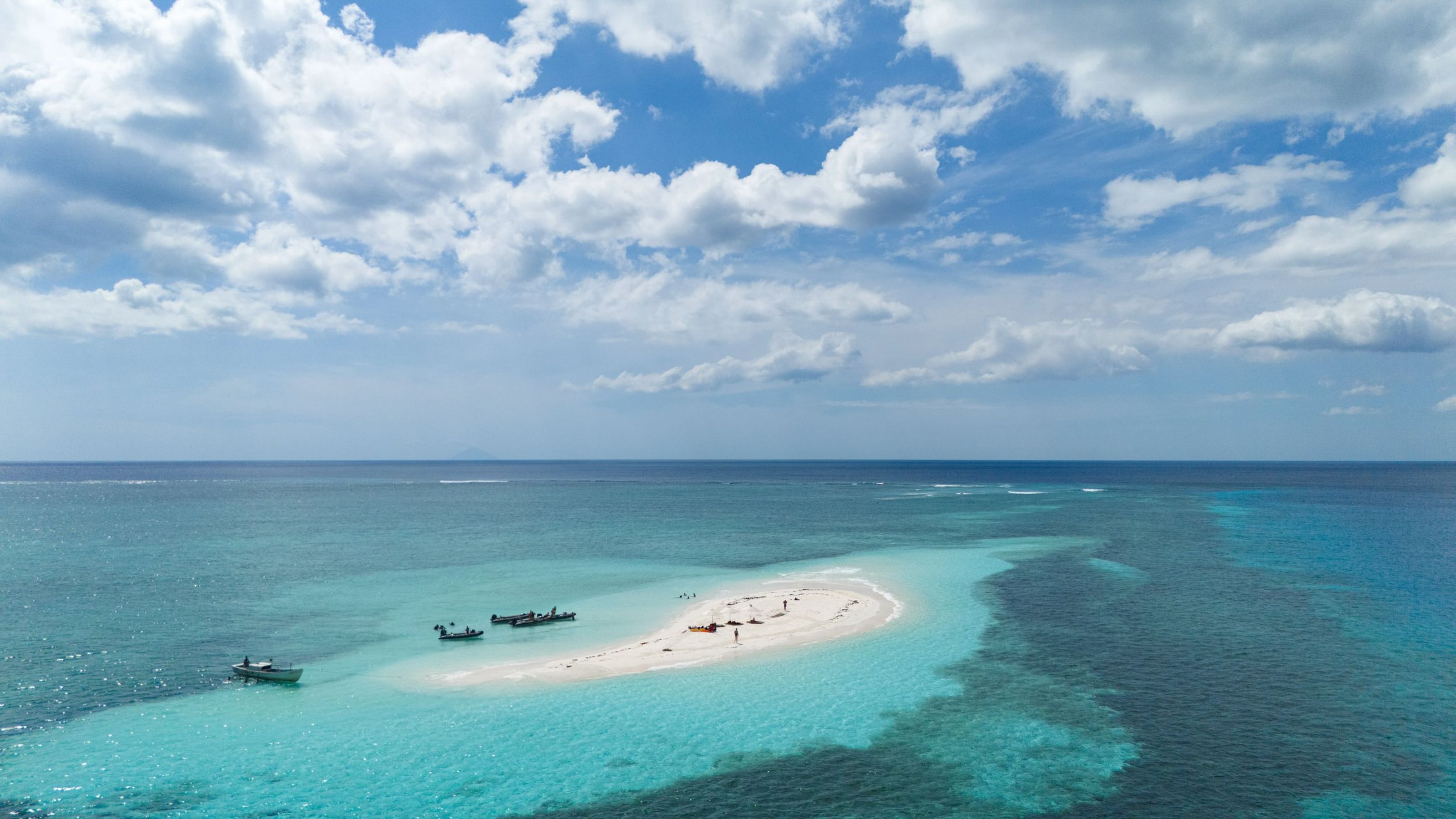 A white sandbar ringed by reef with boats nearby, from the air