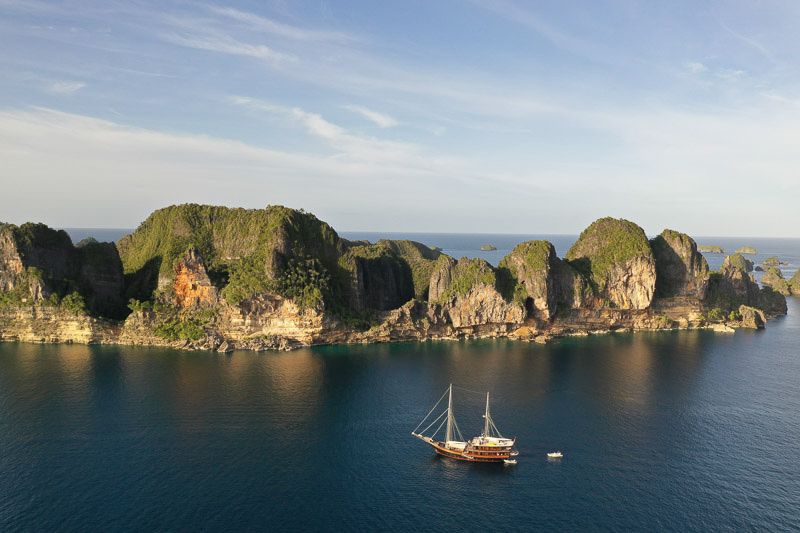 A phinisi at anchor below forested limestone cliffs