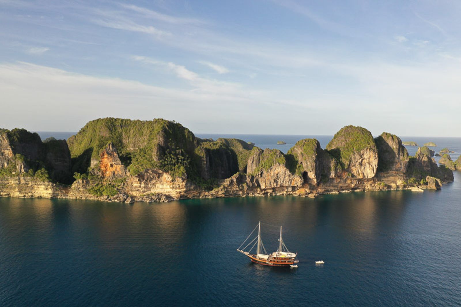 A phinisi at anchor below forested limestone cliffs