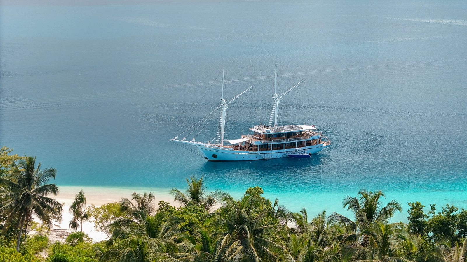 An aerial view of Siloina at anchor beyond palm trees and a white sand beach.