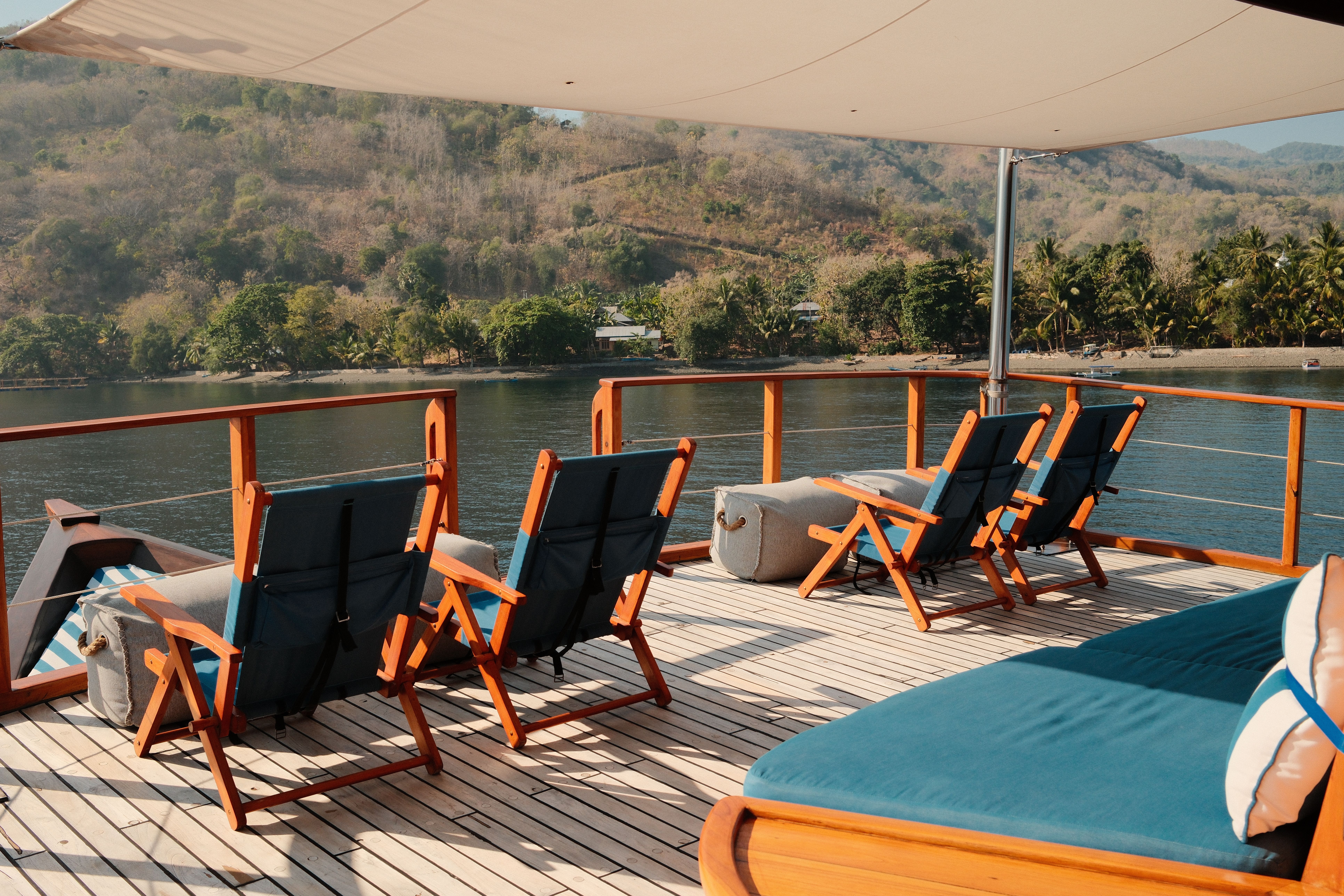 Empty deck chairs and a daybed under the shade awning, a forested shore beyond