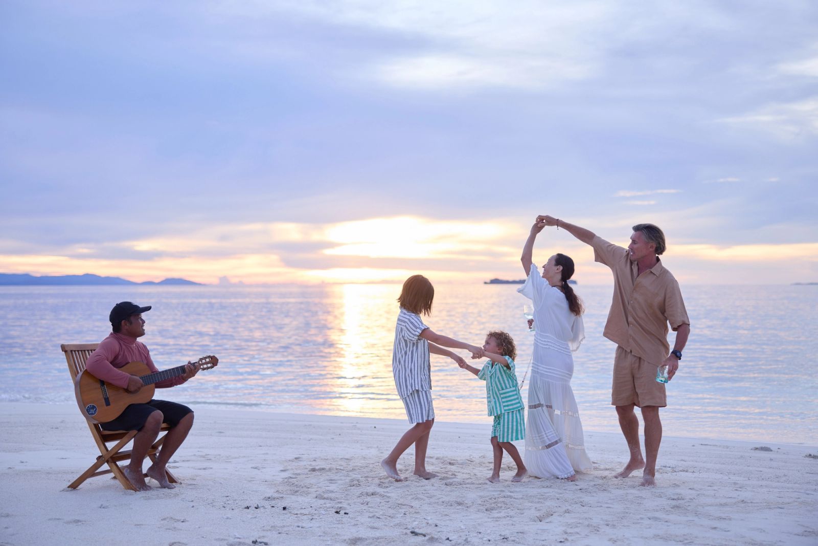 A family dancing on the beach at sunset beside a guitar player