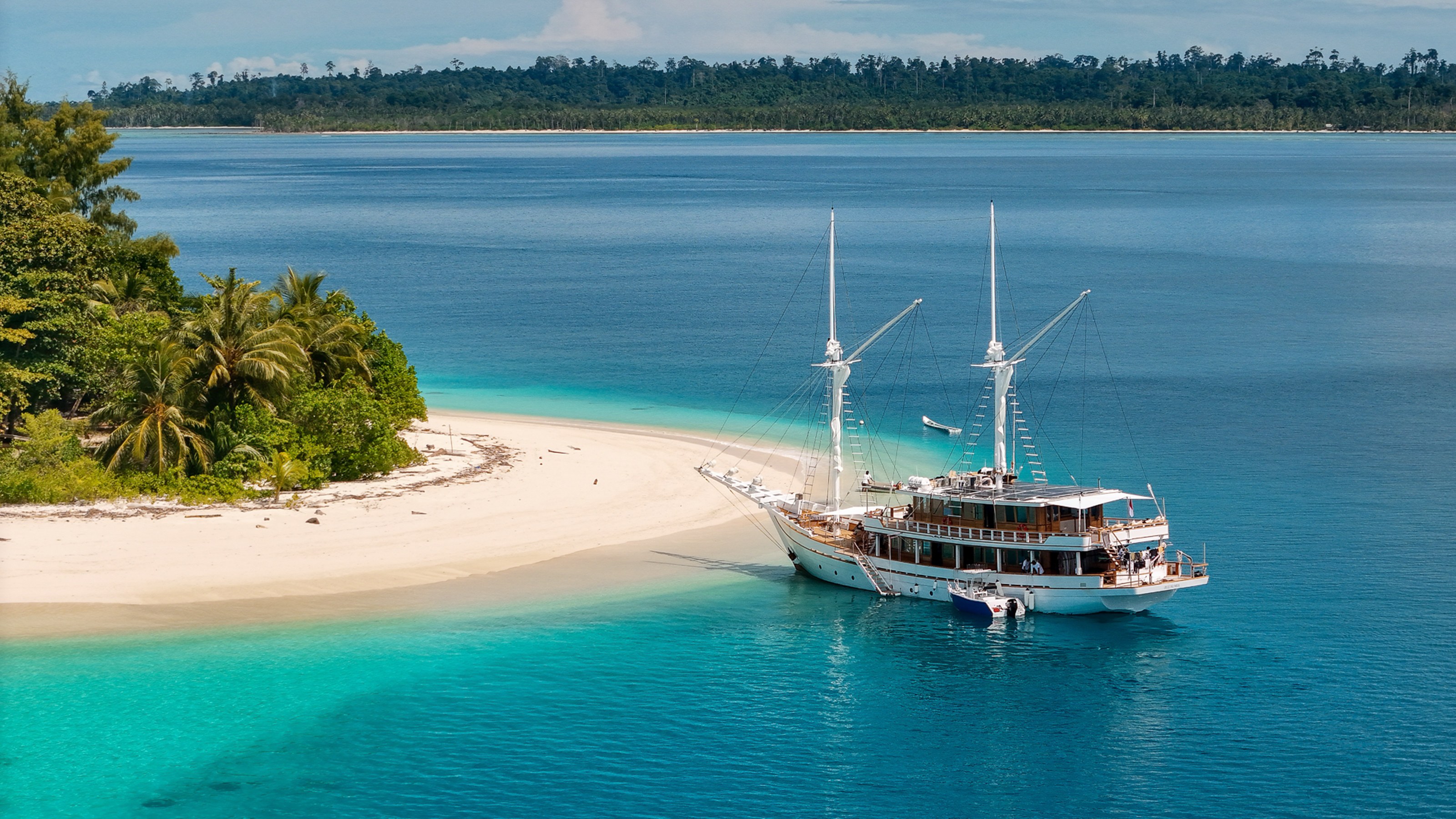Siloina at anchor off a white sand spit with palm trees and turquoise water.