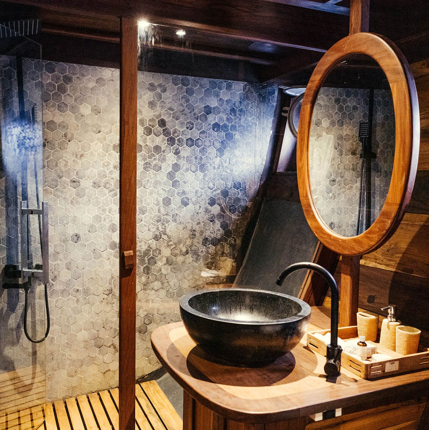 A guest cabin bathroom on Dewata with a black stone basin, a round mirror and a mosaic tiled shower.