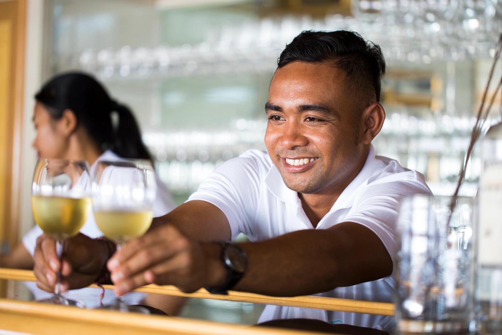 A smiling crew member hands two glasses of white wine across the bar.