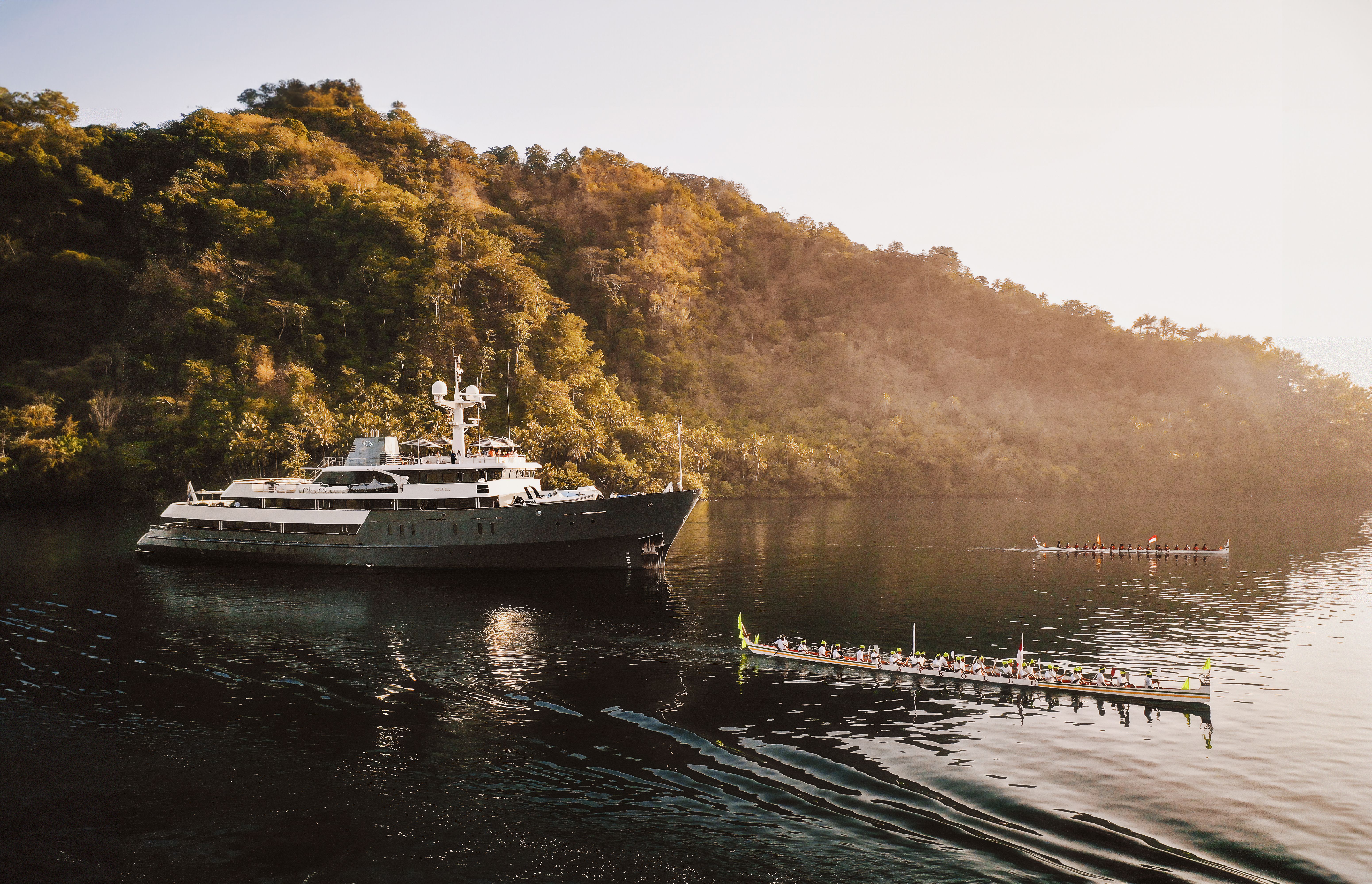 Aqua Blu at anchor beneath forested hills in the Banda Islands as a traditional kora kora canoe rows past in golden light.