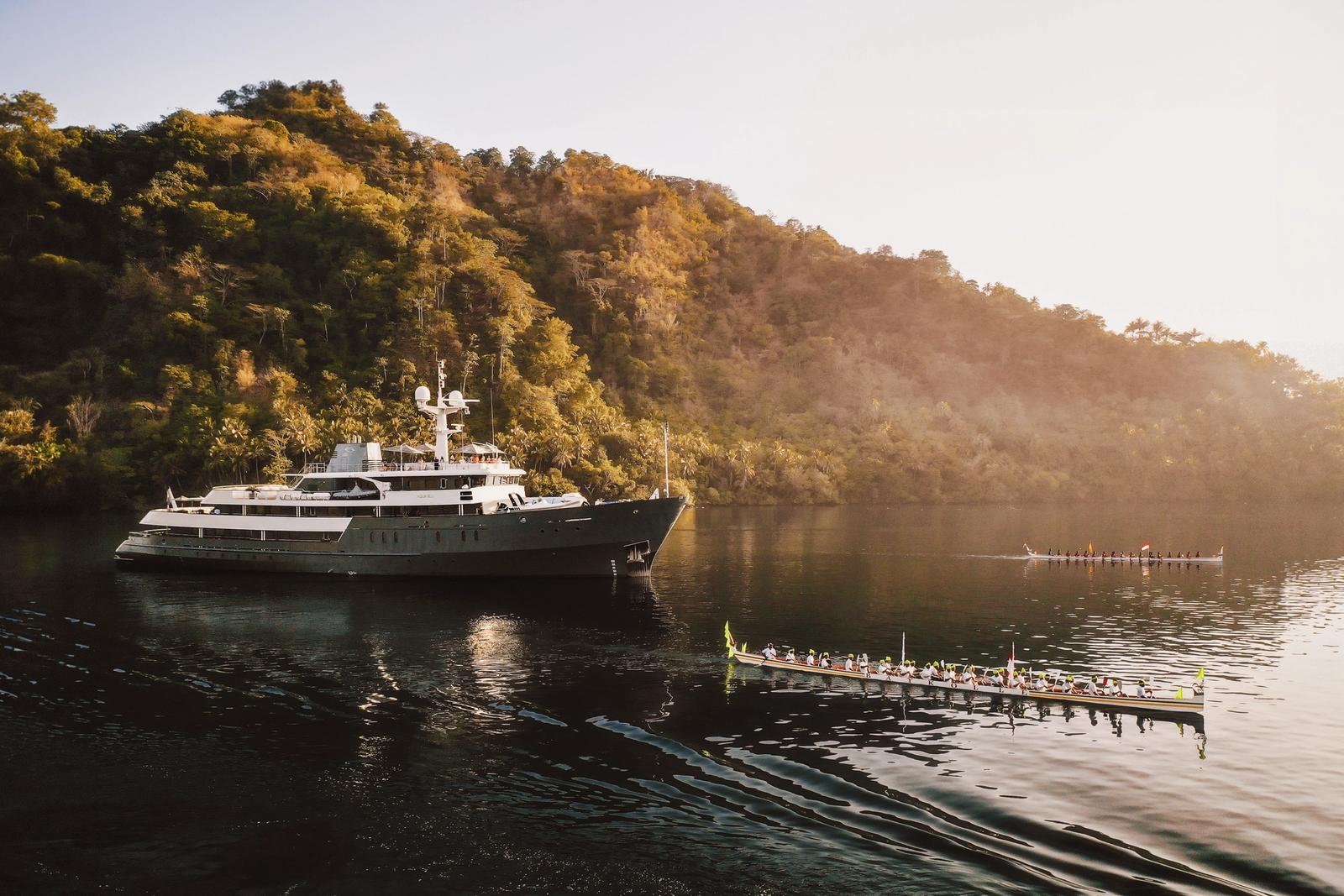 Aqua Blu at anchor beneath forested hills in the Banda Islands as a traditional kora kora canoe rows past in golden light.