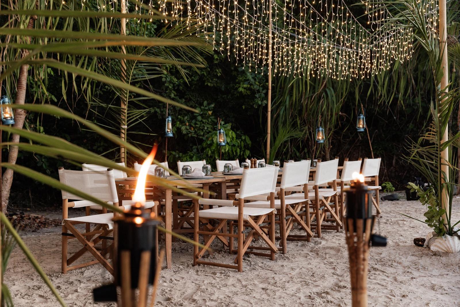 A long dinner table on the sand under trees and lights, with torches
