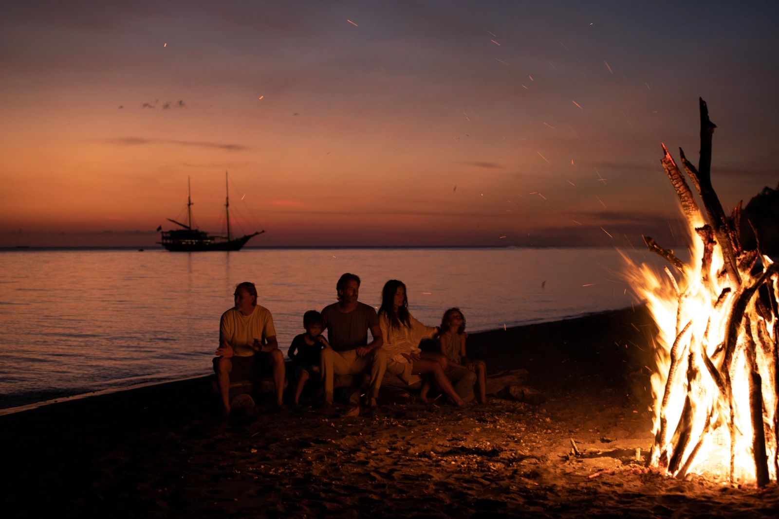 Guests around a bonfire on the beach at dusk, a phinisi offshore