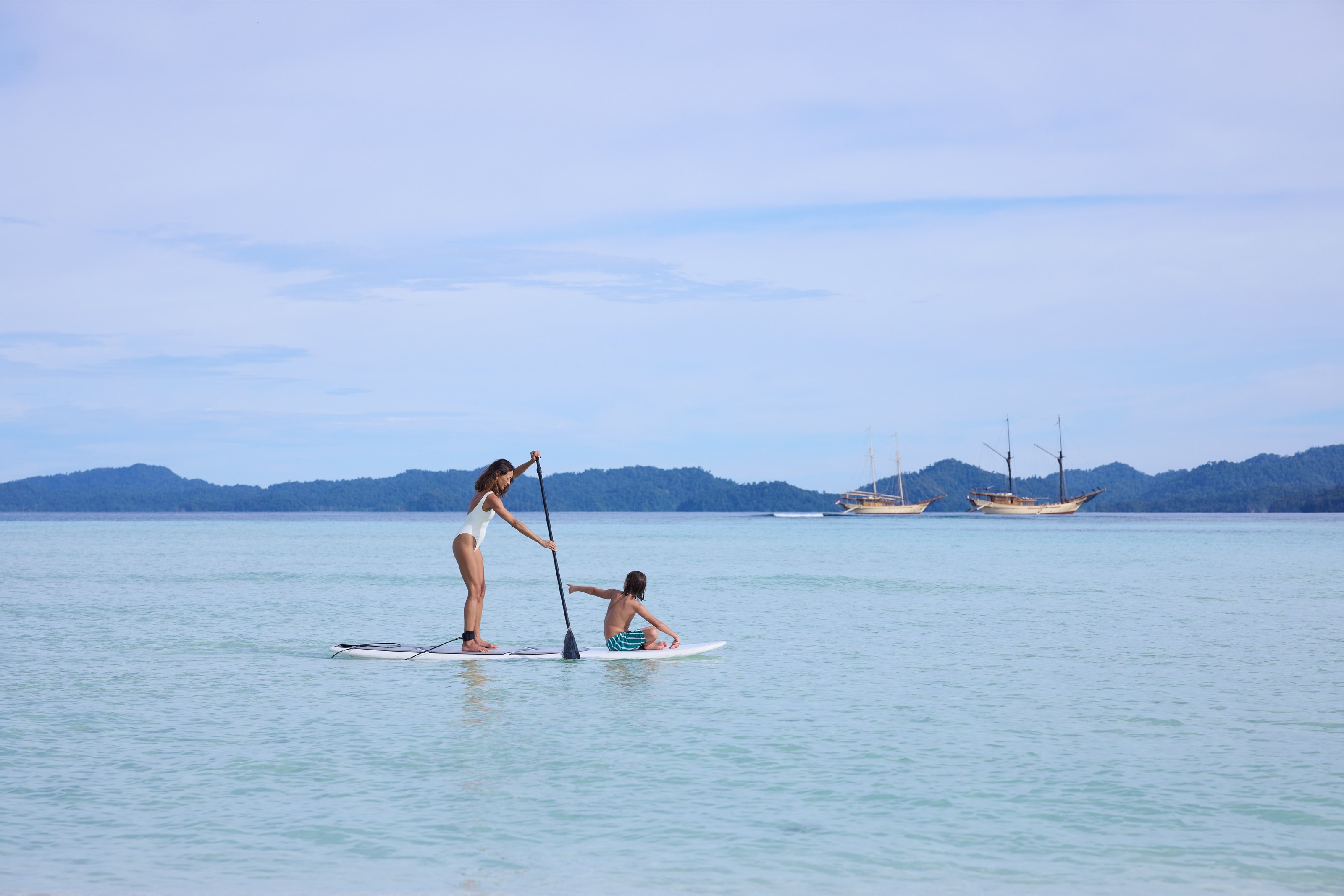 A mother and son on a paddleboard in calm shallows, two phinisis anchored in the distance