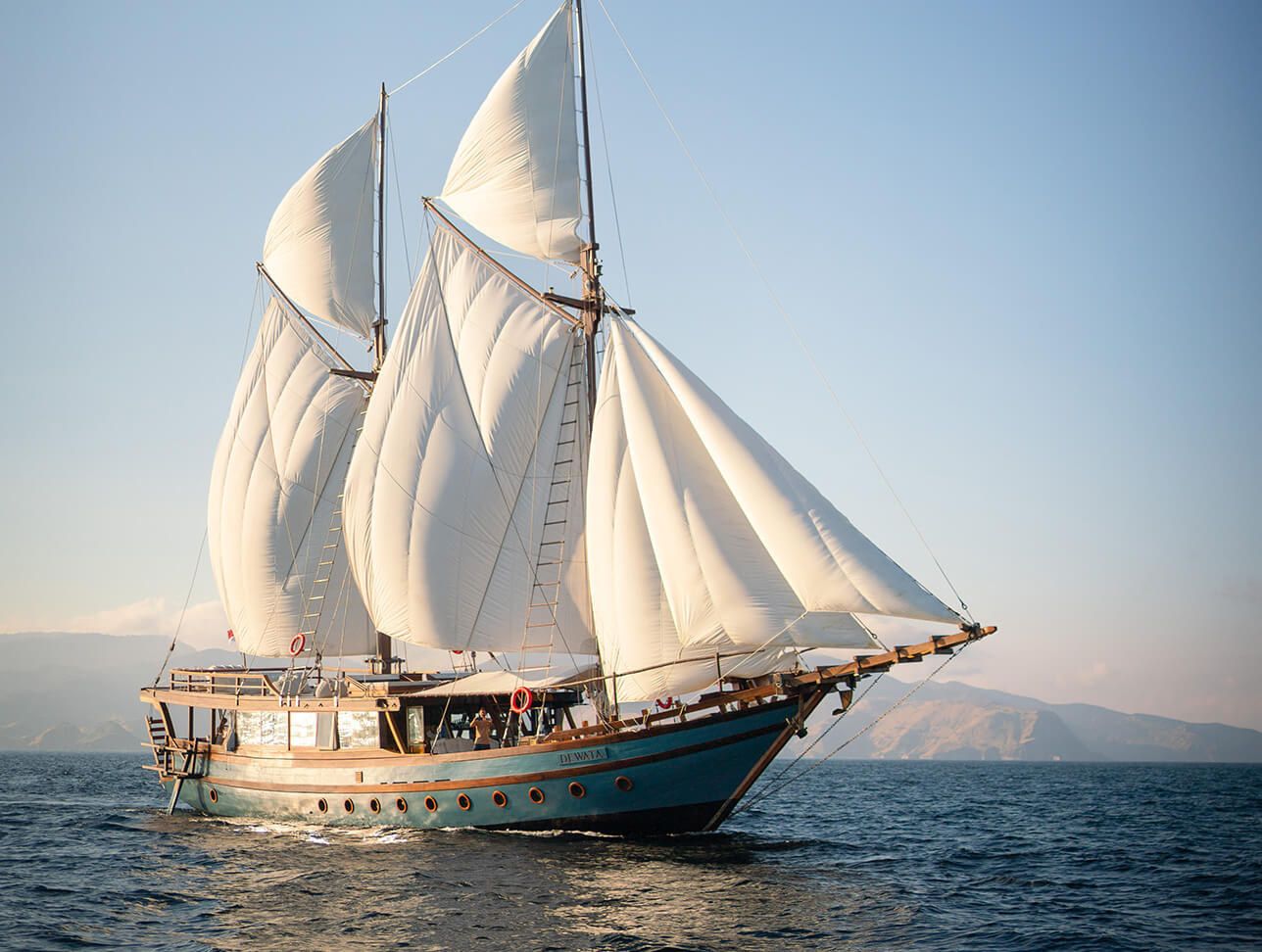 A phinisi under full sail in golden light with hills on the horizon.