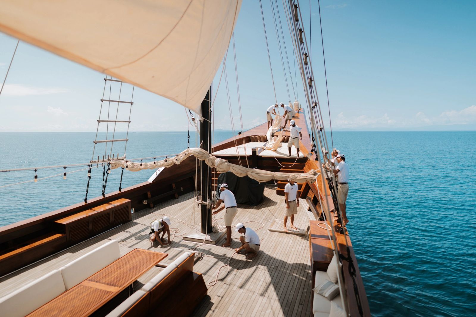 The deck of Celestia running forward beneath a cream sail with crew at work.