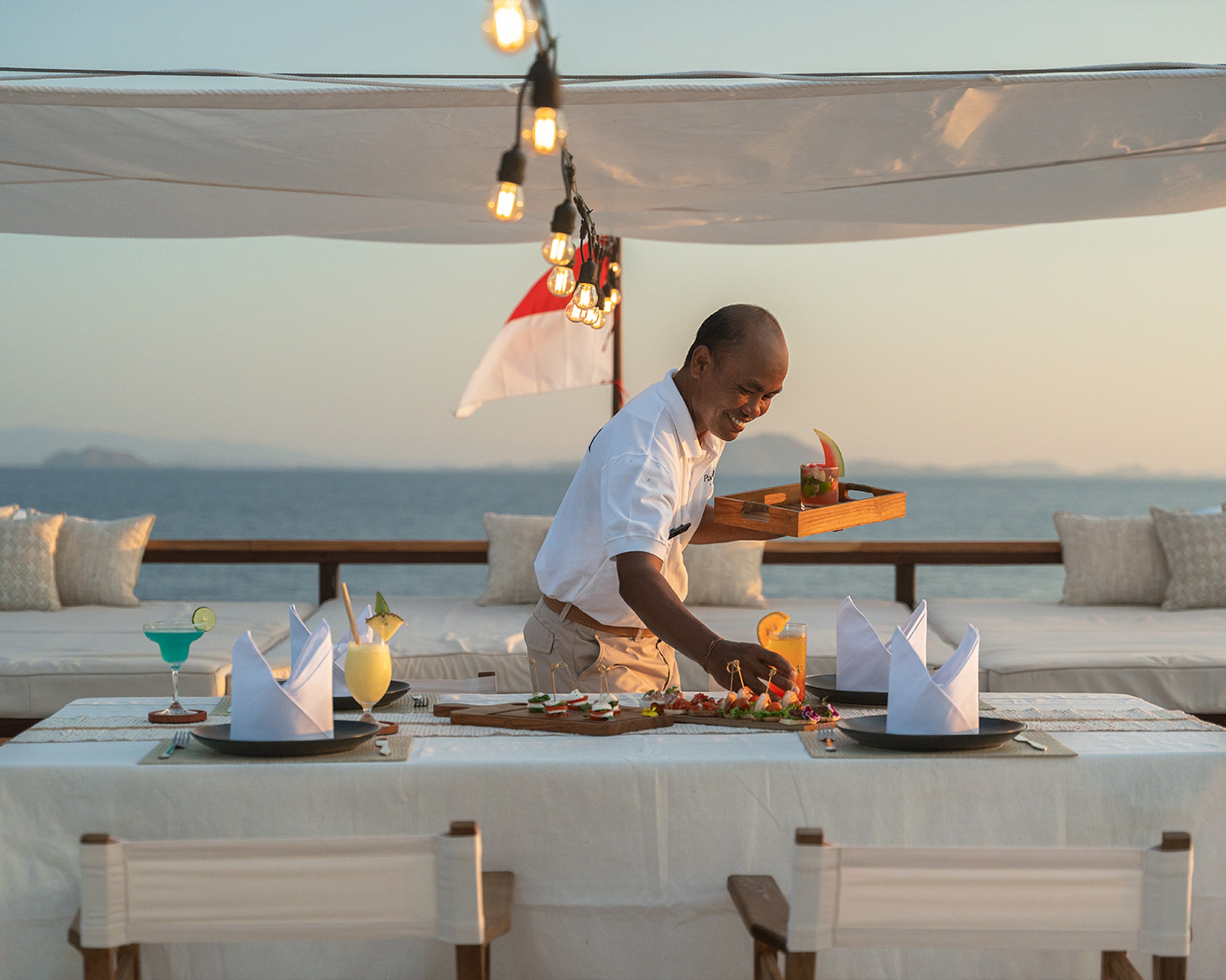 A crew member arranges food on the laid dining table on the deck of Sanya at sunset.