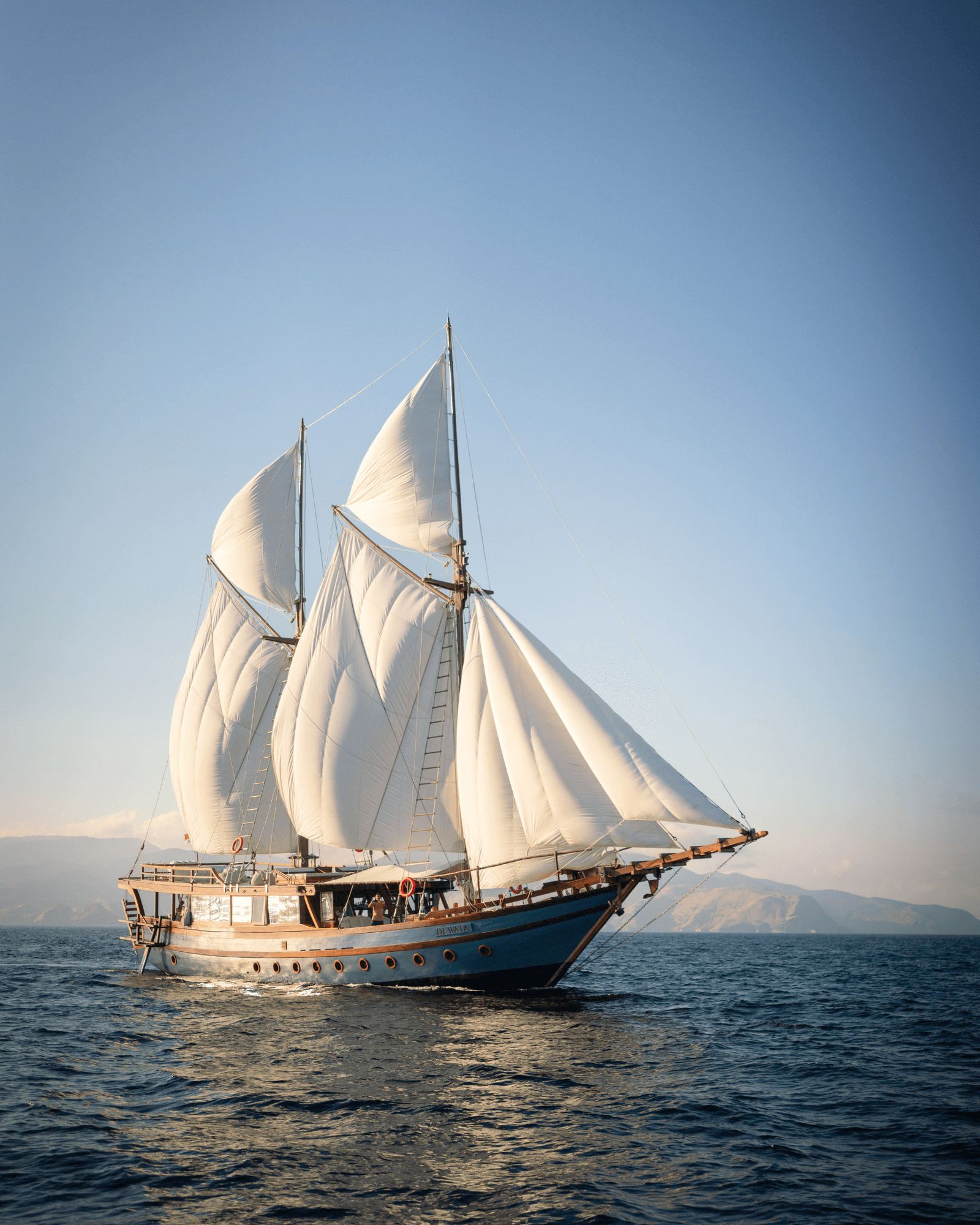 A phinisi with a white hull and white sails set sailing on a calm sea.