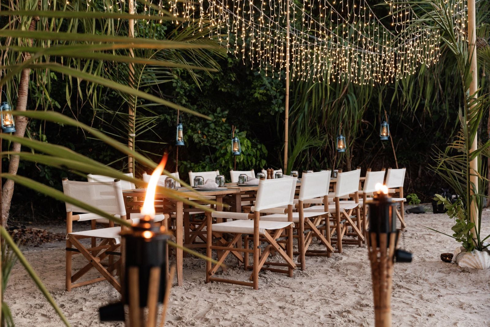 A long dinner table on the sand under trees and lights, with torches