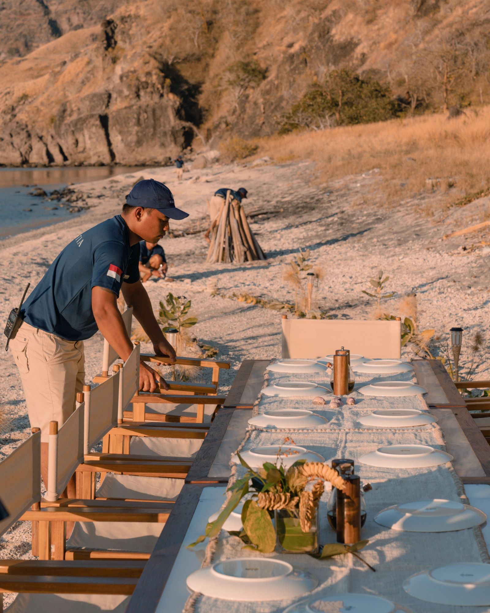 A crew member setting the long beach dinner table beside the rocks.