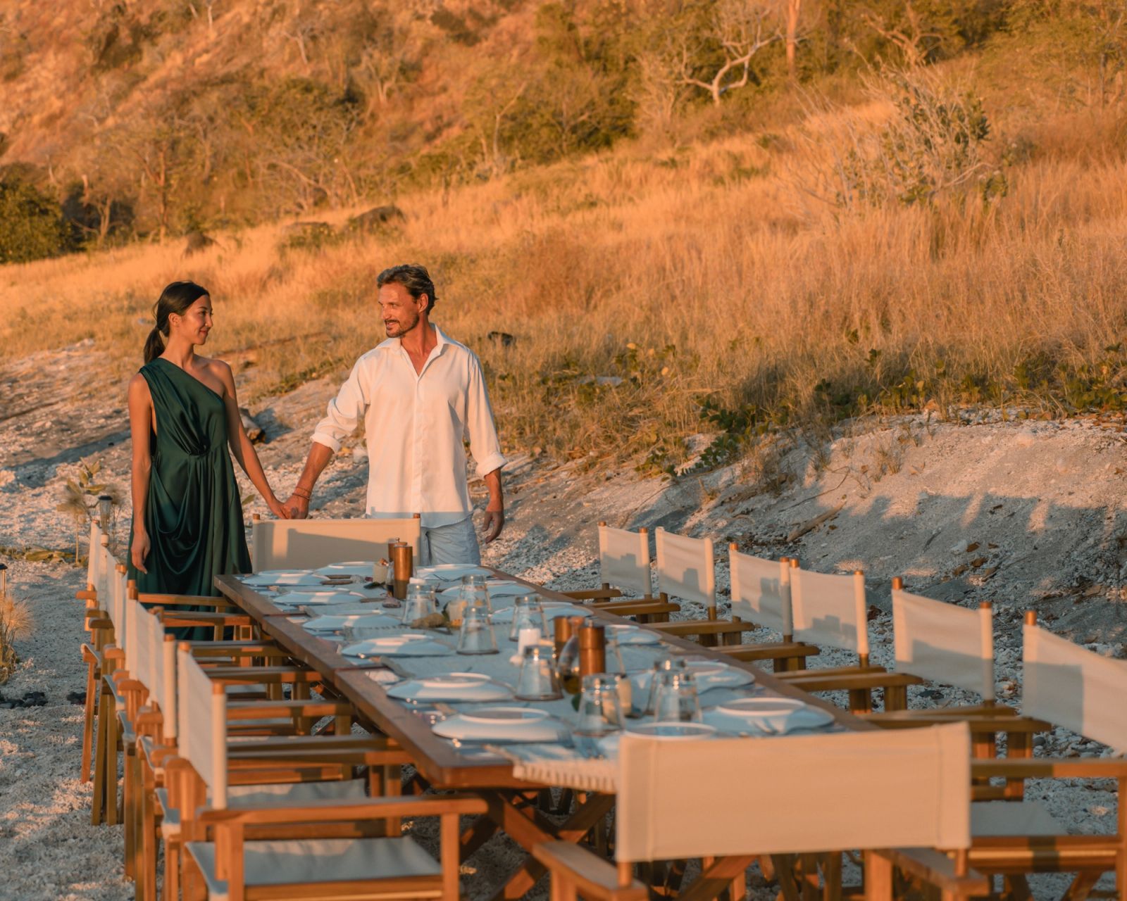 A couple walking hand in hand beside the long beach dinner table at sunset.