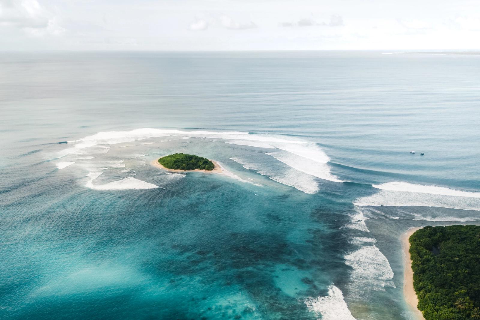 Waves wrapping a small island and its reef pass in the Telo Islands, from the air