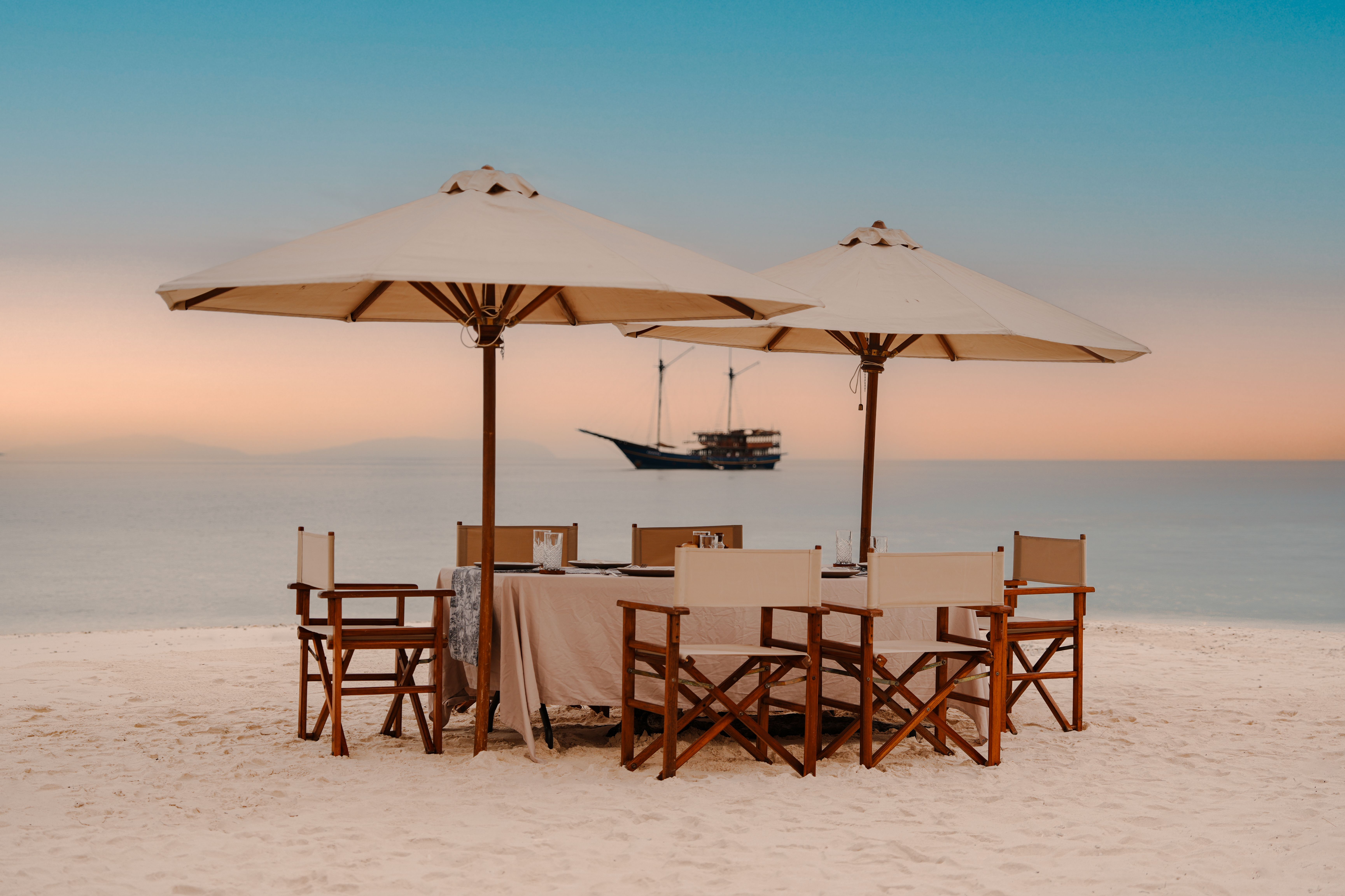 A dinner table under two parasols on a white-sand beach at dusk, with a phinisi anchored offshore.