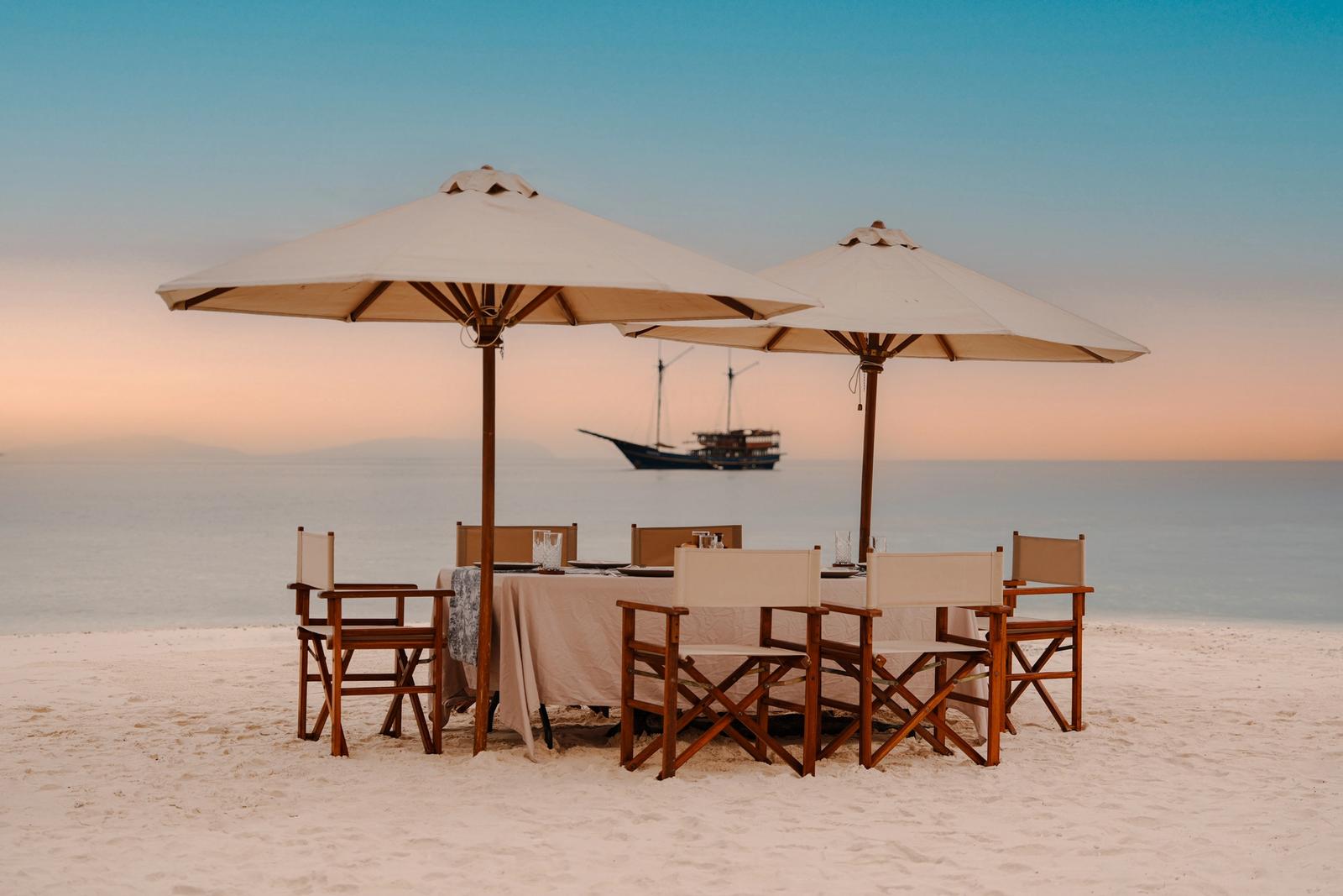 A dinner table under two parasols on a white-sand beach at dusk, with a phinisi anchored offshore.