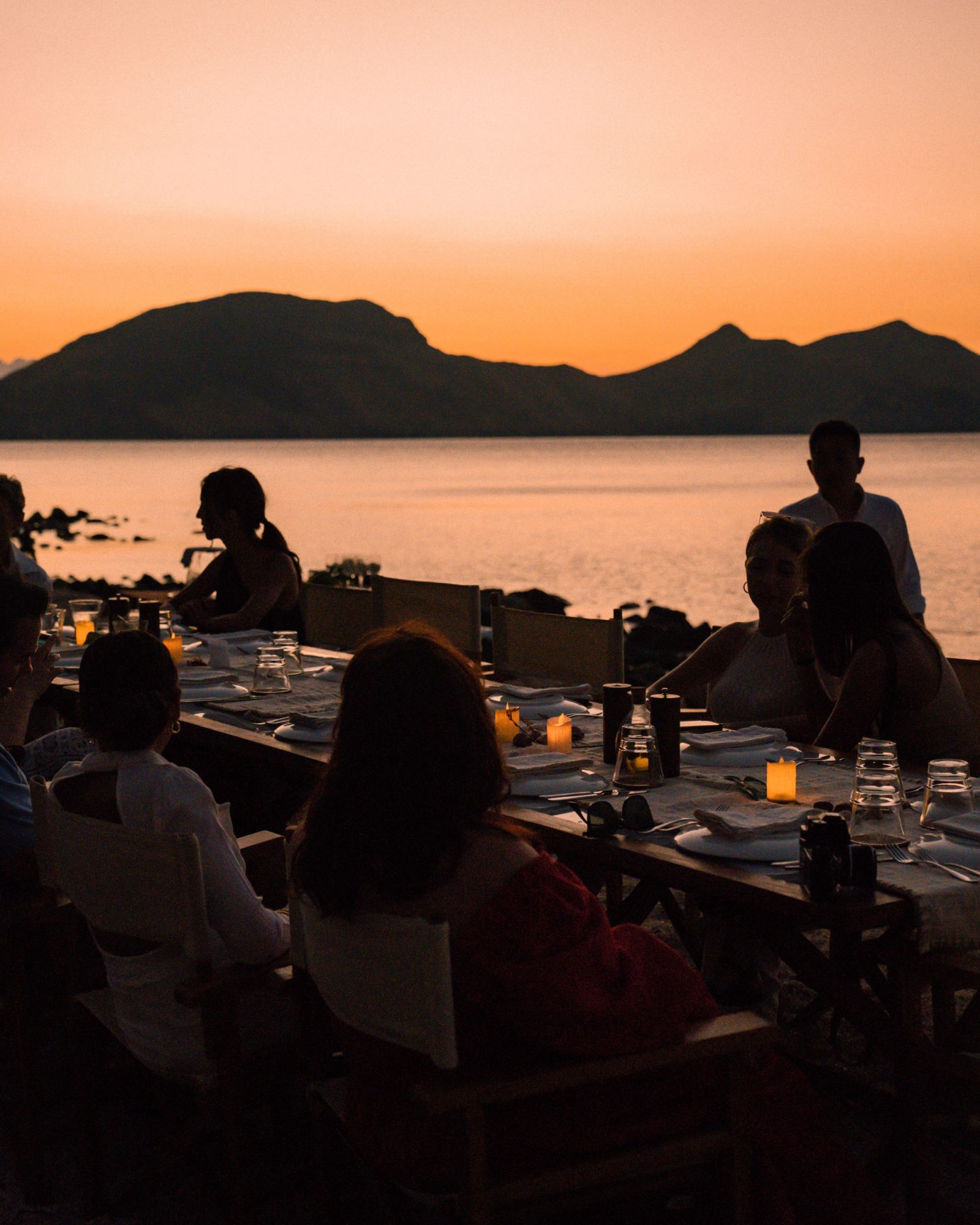 Guests seated at a candlelit beach dinner at sunset.