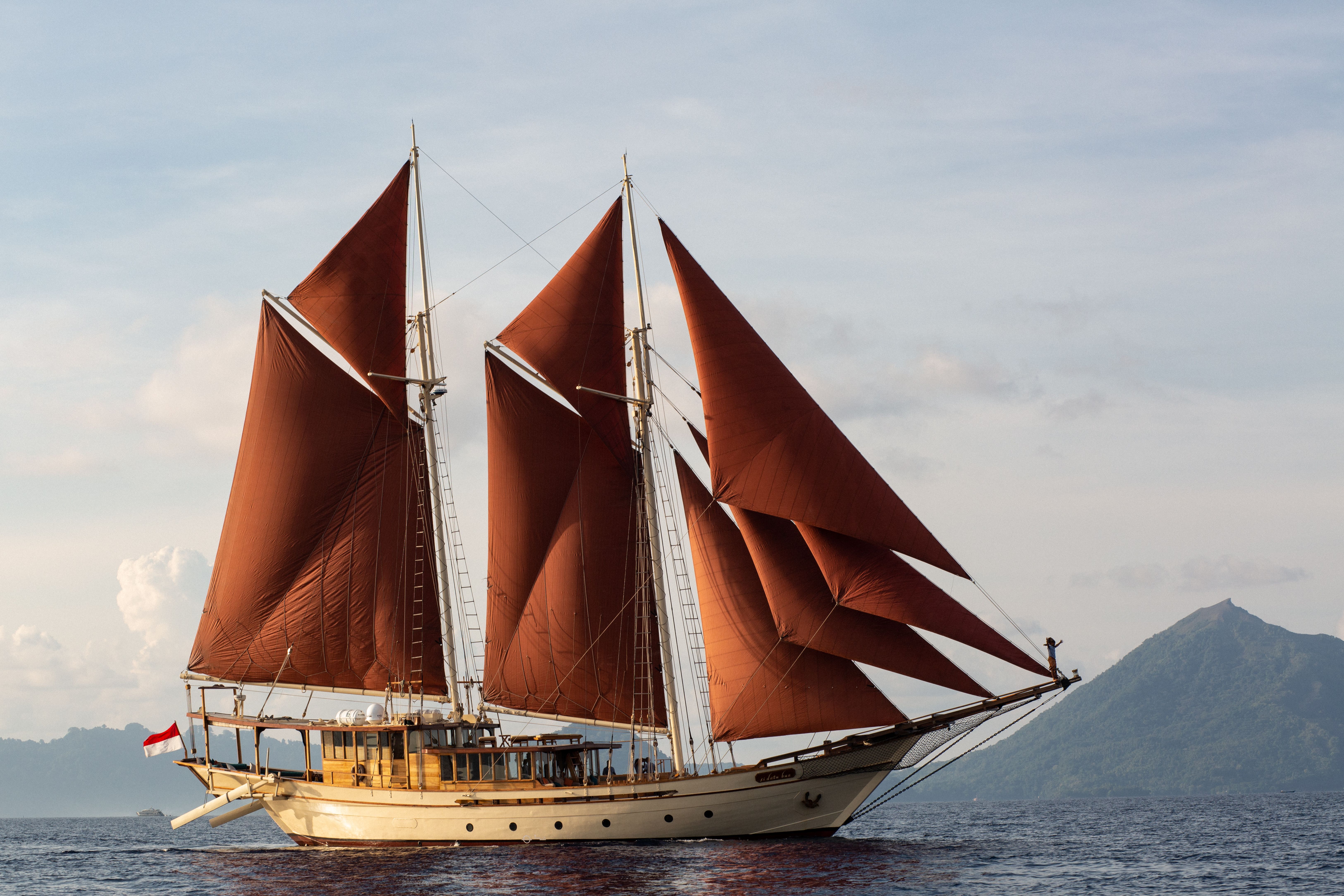 Si Datu Bua sailing under her red sails off a volcanic coastline.