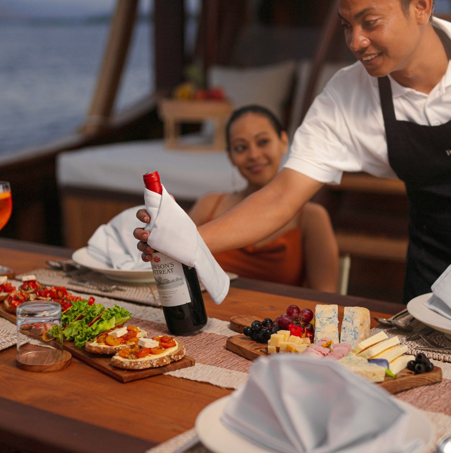 A crew member pouring wine for a guest at a table laid with seafood and cheese.