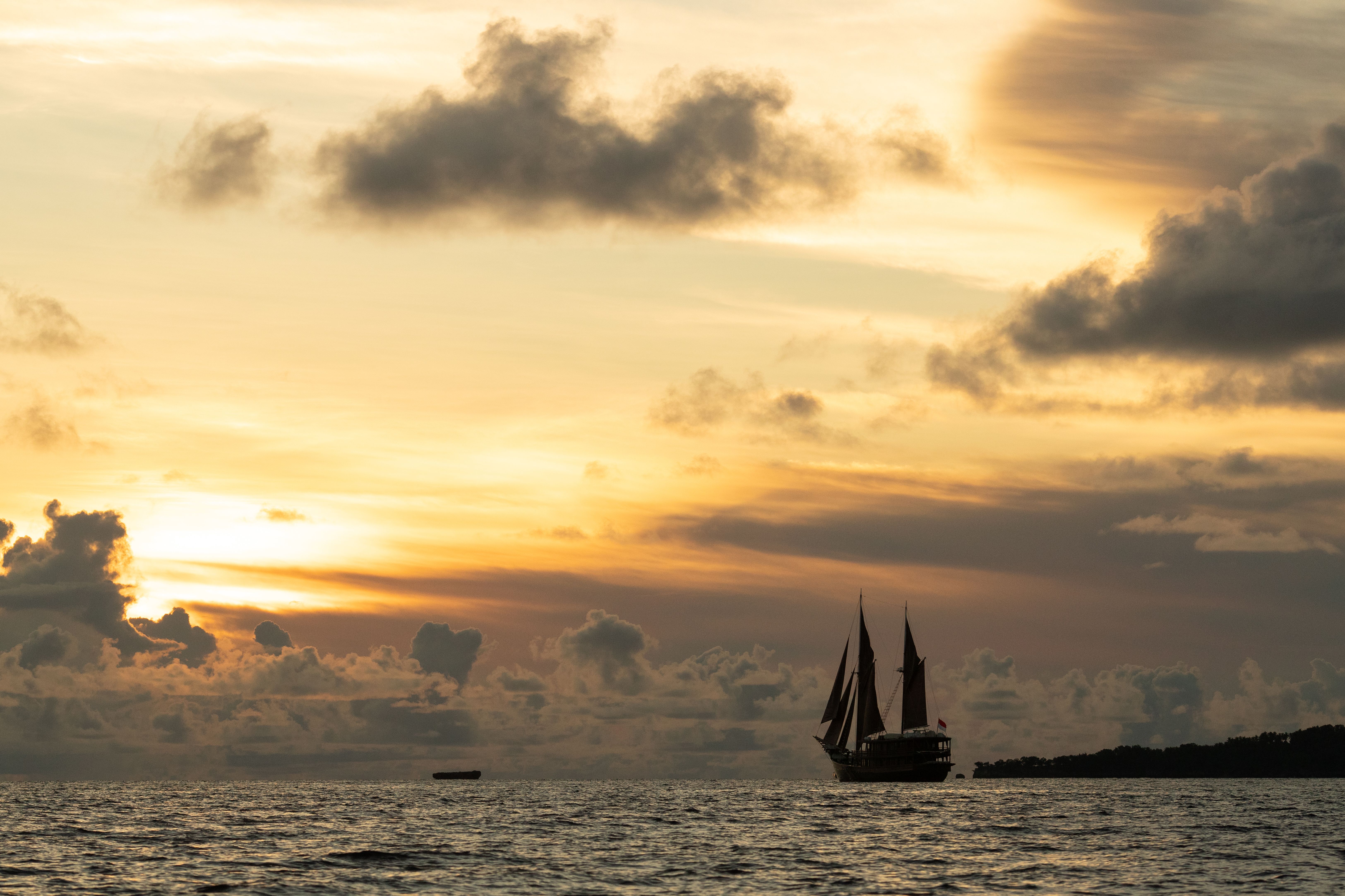 Dunia Baru silhouetted under sail against a golden sunset sky.
