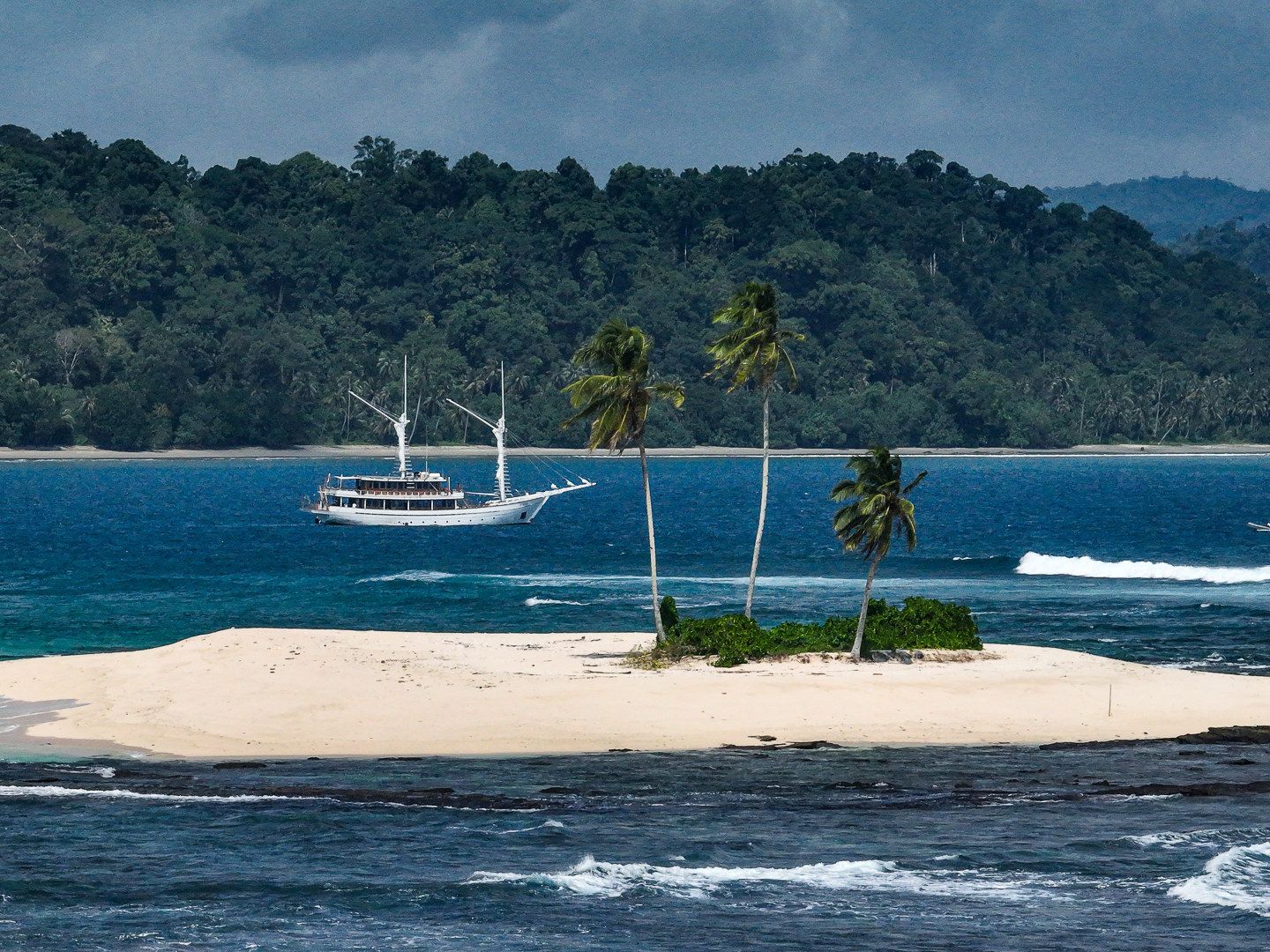 Siloina anchored off a white sand islet with palm trees and forested hills behind.