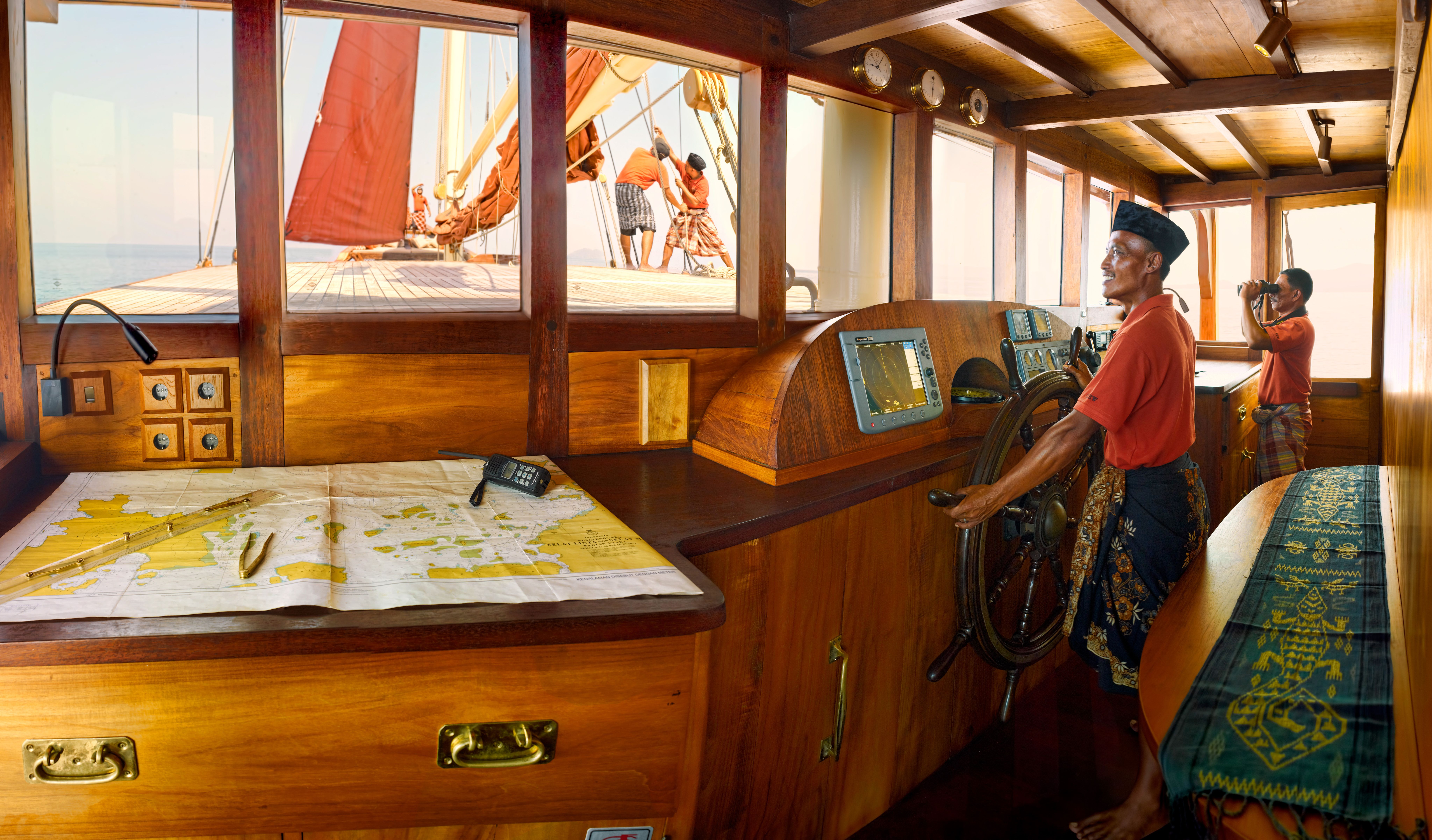 The captain at the wheel of a phinisi's wooden wheelhouse, a nautical chart spread on the table and crew working the sails outside.