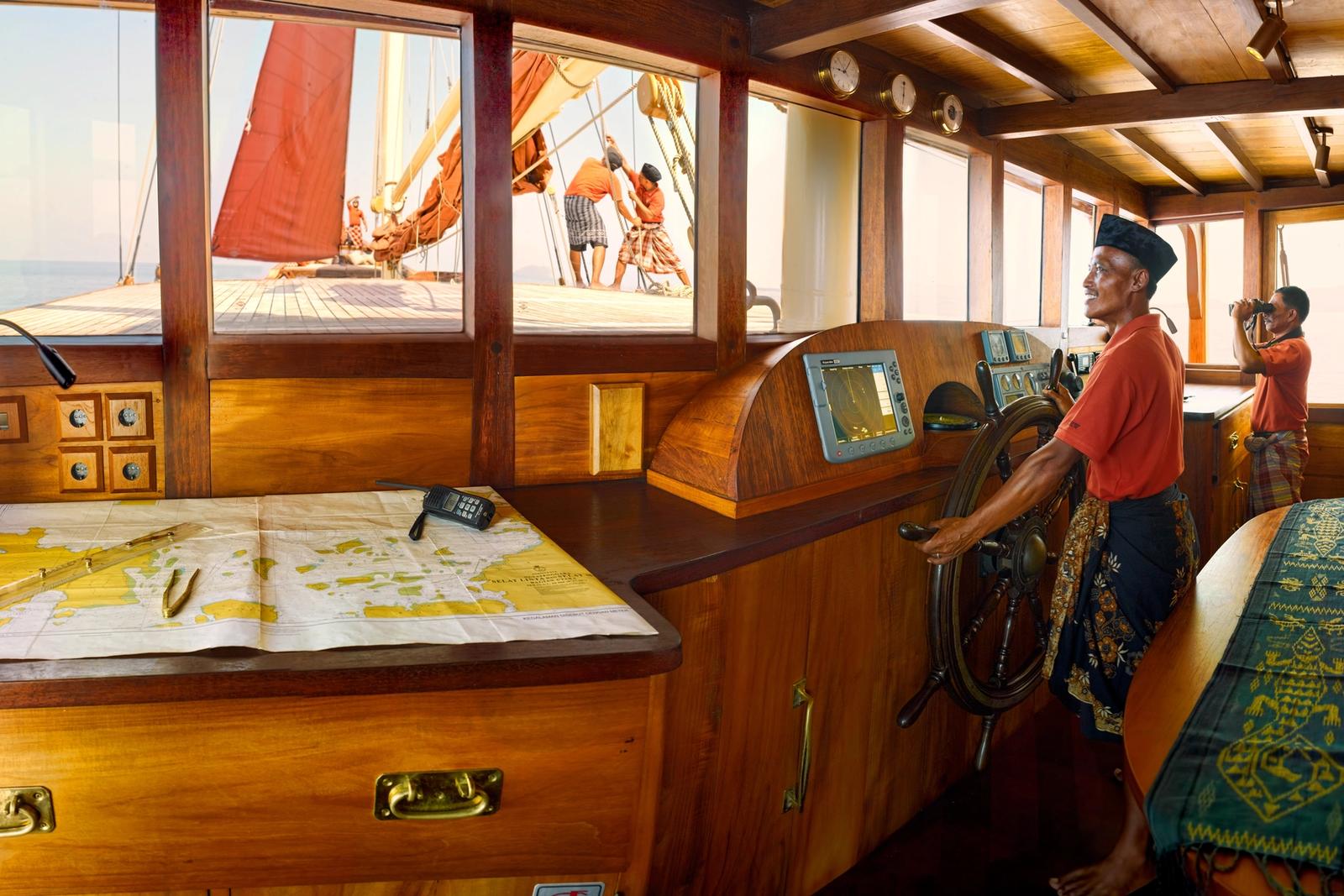 The captain at the wheel of a phinisi's wooden wheelhouse, a nautical chart spread on the table and crew working the sails outside.