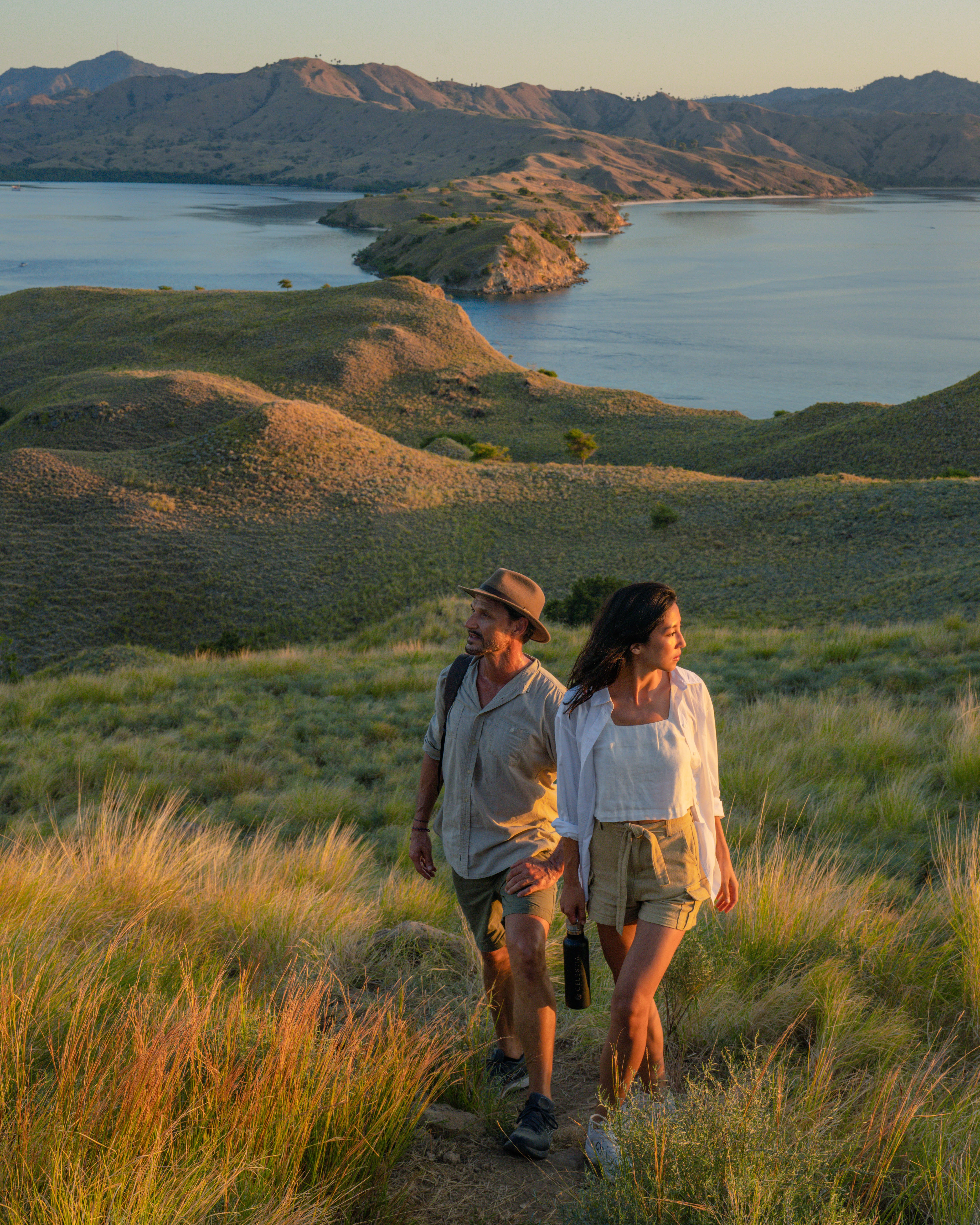 A couple walks a grassy ridge above the bays of Komodo National Park in evening light.