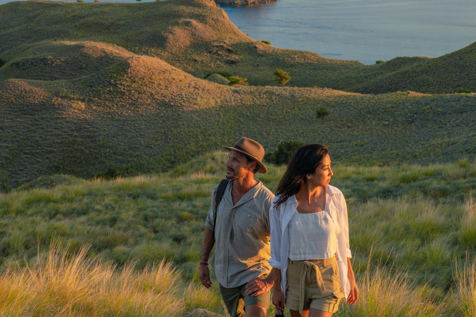 A couple walks a grassy ridge above the bays of Komodo National Park in evening light.