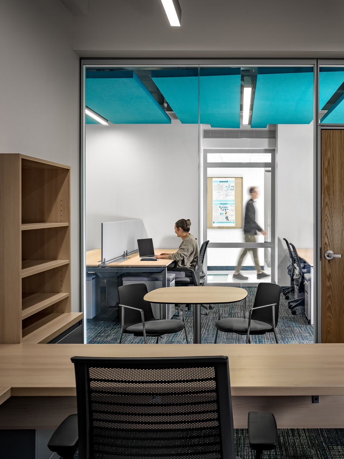 View from faculty office into the open work area where a woman is working at a desk and a person can be seen walking in the hallway through a window